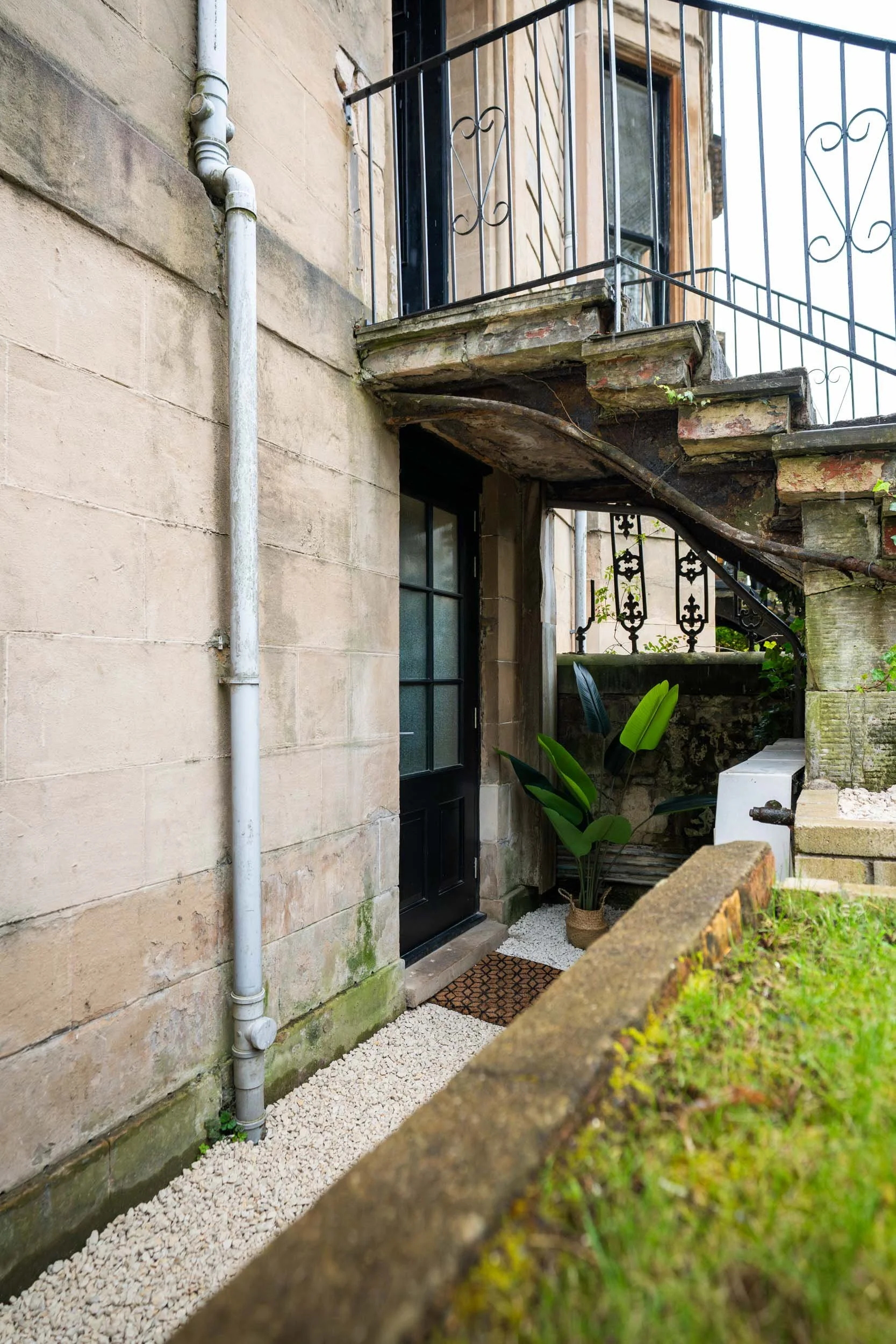 Exterior view of an old building with a black door, stone walls, a small porch, and a metal staircase leading to an upper balcony. A potted plant is next to the door, and the ground is covered in white gravel.