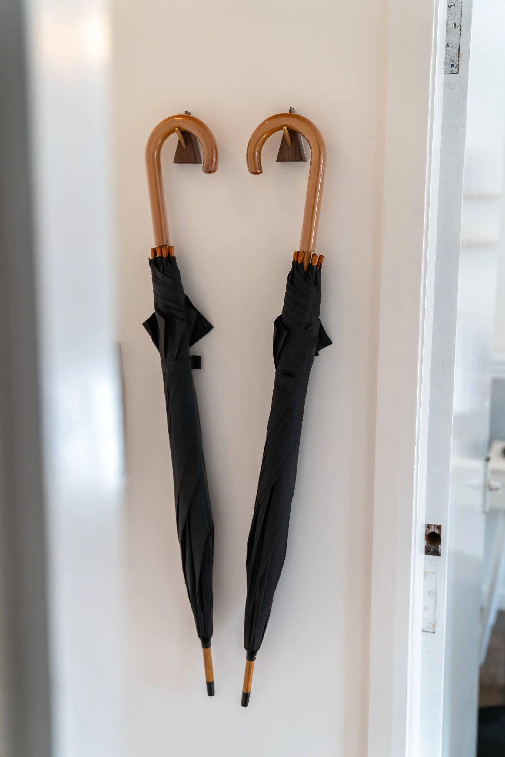 Two umbrellas hanging on a white wall, one with a natural wooden curved handle and the other with a similar handle, both wrapped in black covers.