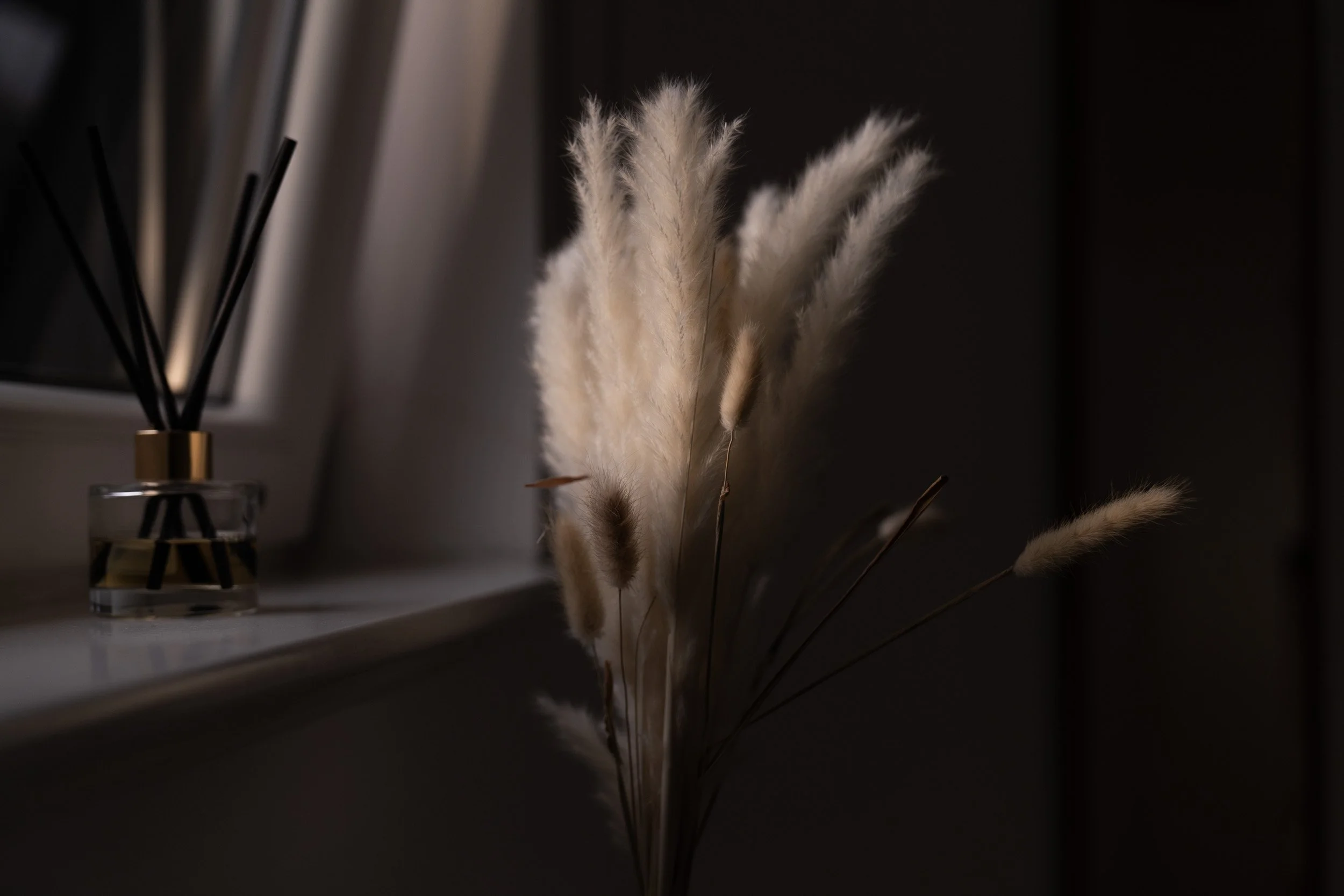 A bunch of fluffy beige and white dried pampas grass in a dark room, with a container of black reed diffuser sticks on a windowsill.