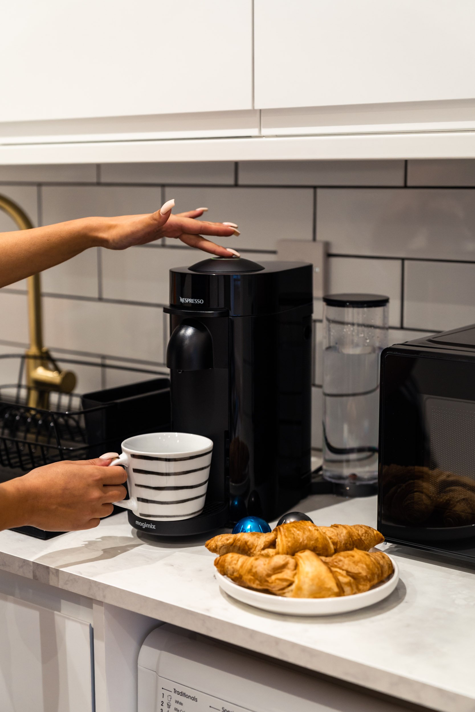 A person is pouring coffee into a striped mug using a black Nespresso coffee machine. There are croissants on a white plate and a microwave on the countertop in a modern kitchen with white cabinets and white subway tile backsplash.