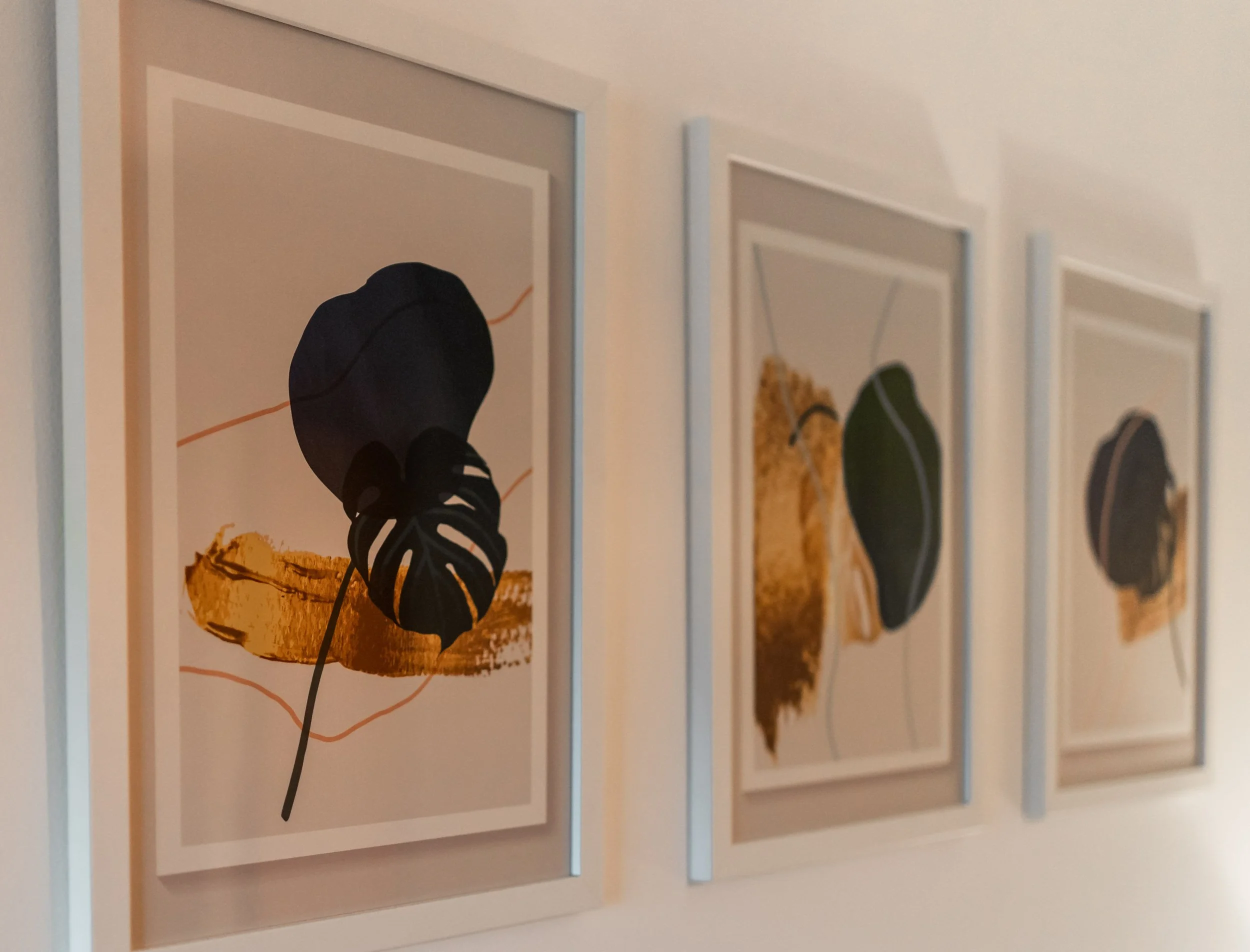 Three framed art prints on a wall, each featuring abstract botanical shapes with black leaves, gold accents, and minimalistic line details.