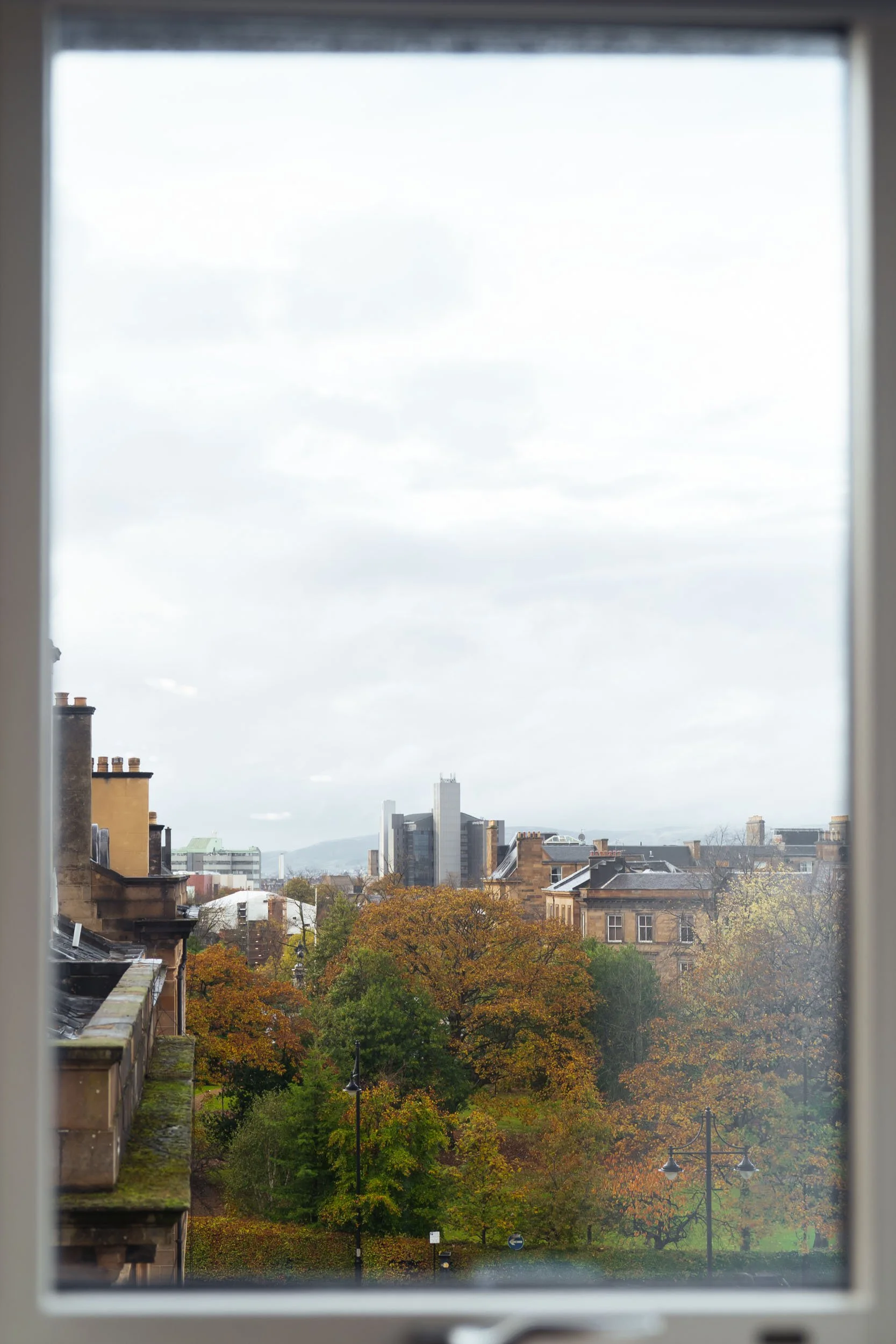 View through a window showing a cityscape with trees in autumn and tall modern buildings in the distance.
