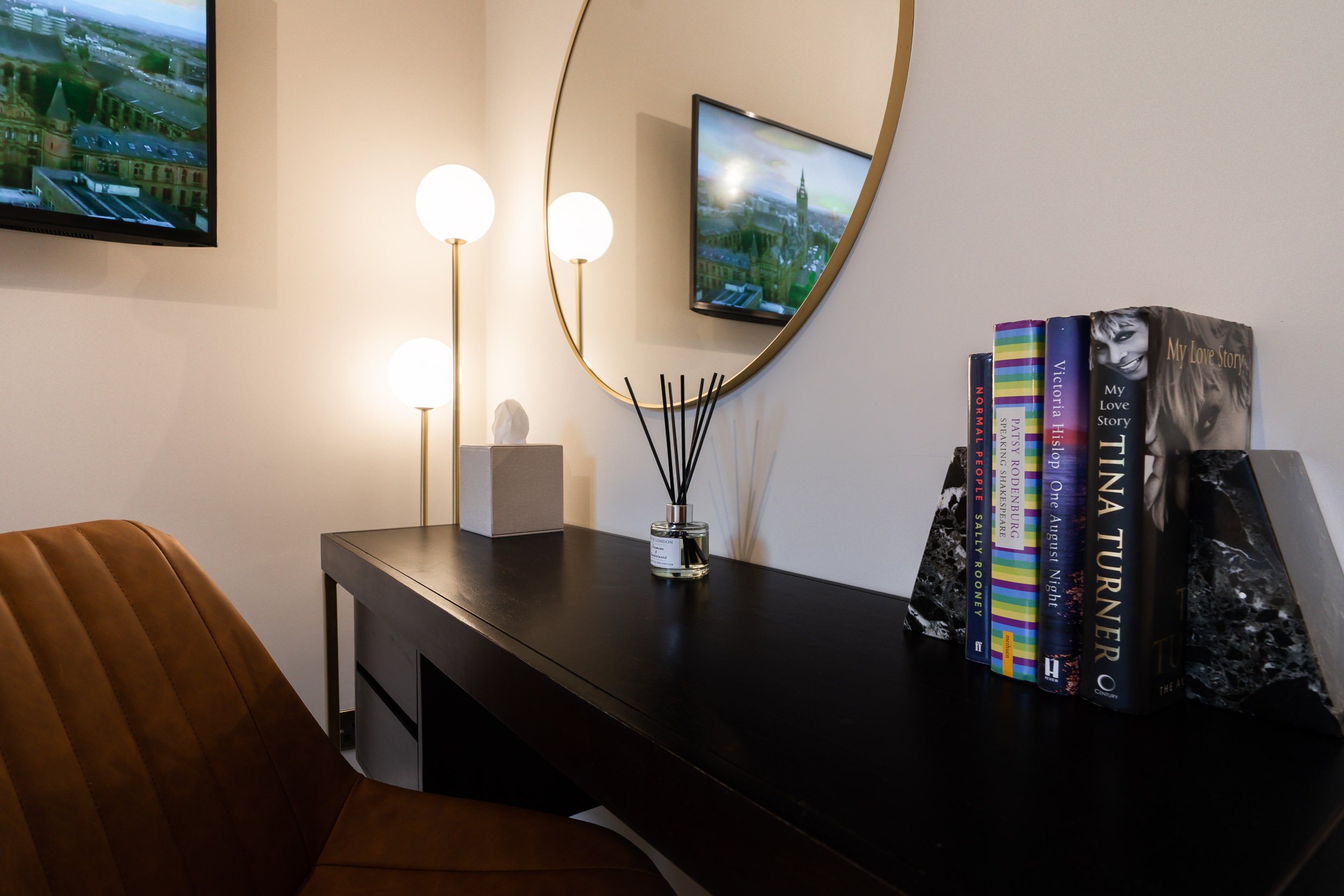 A corner of a room with a desk, a brown chair, a wall-mounted TV, a round mirror, a three-globe floor lamp, a box with a sculpture, an incense diffuser, and a row of books on black and marble bookends.