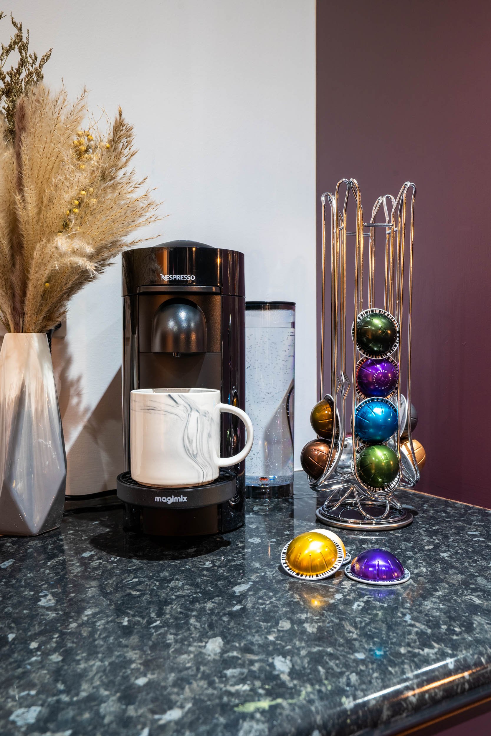 Coffee station with a Nespresso machine, a white mug on a black Magimix tray, a water carafe, and a holder of colorful Christmas ornaments on a dark granite countertop.