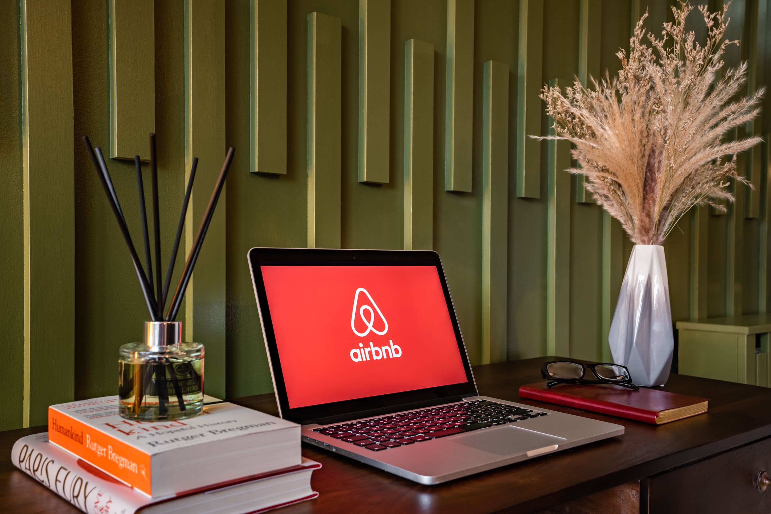 Desk with books, a laptop displaying the Airbnb logo, glasses, a red notebook, and vases with dried plants against a green wall with vertical panels.