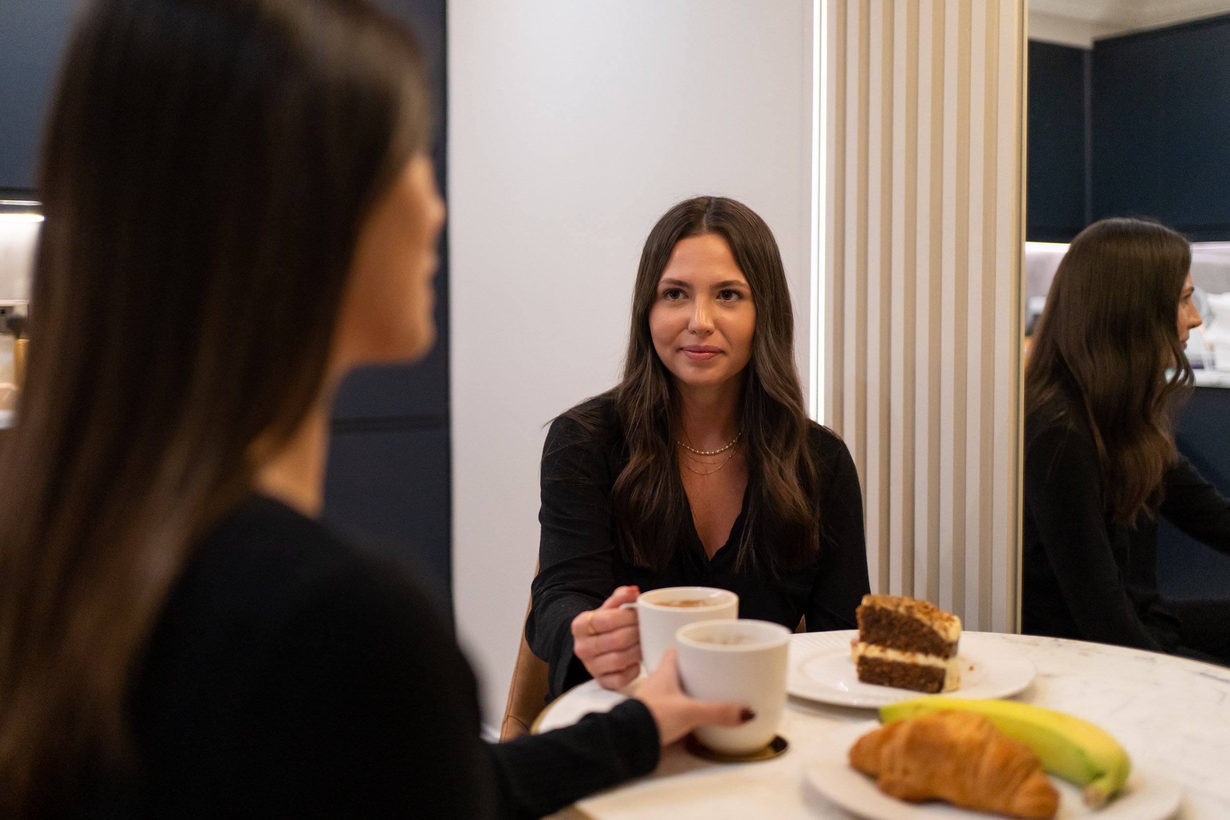 Two women sitting at a table having coffee, with cake and bananas on the table, one facing the camera and holding a coffee cup, the other with her back to the camera.