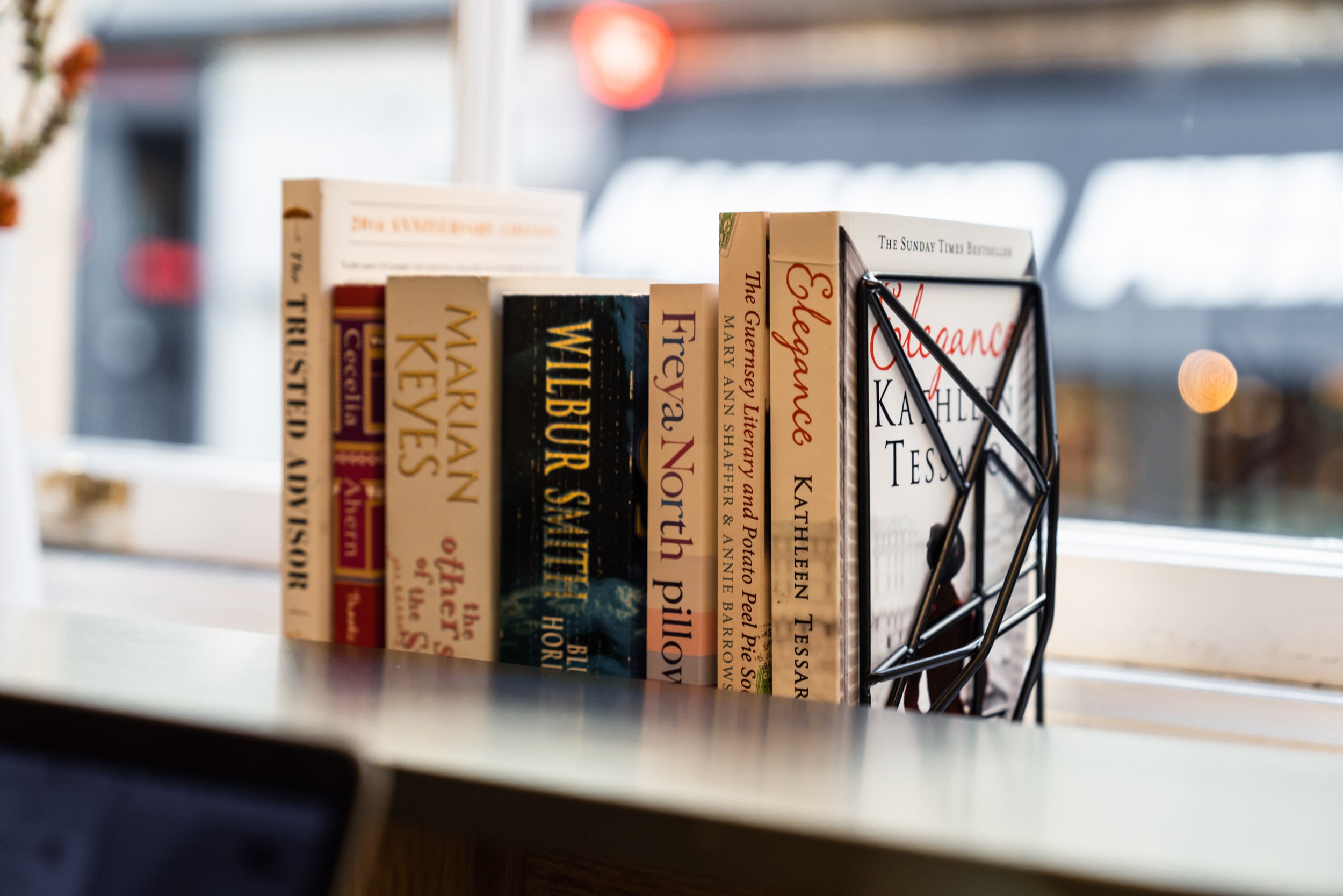 A collection of books in a black wire book holder on a windowsill, with titles including "Weber Steward," "Marian Keys," "Freya North," and "Kathleen Tessaro."