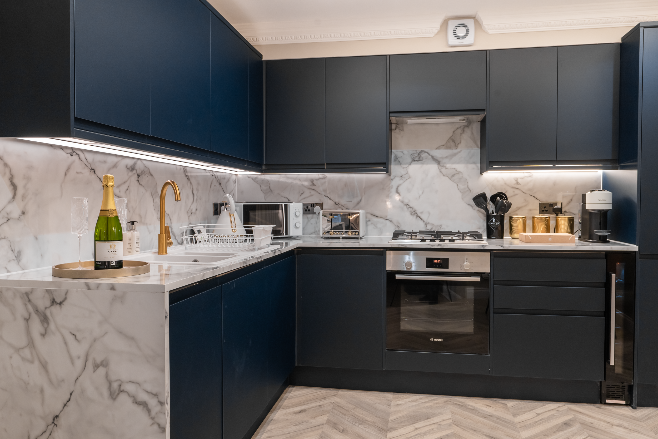 Modern kitchen with dark blue cabinets, white marble backsplash, gold faucet, small appliances, and wine bottle with glasses on the counter.