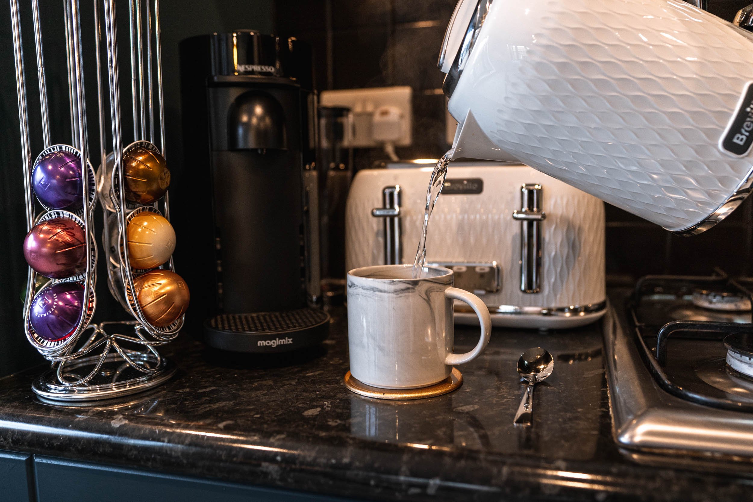 Coffee being poured into a mug from a white pitcher on a black kitchen countertop, with a spoon and a metallic toaster in the background, and a stand with colorful coffee capsules on the left.