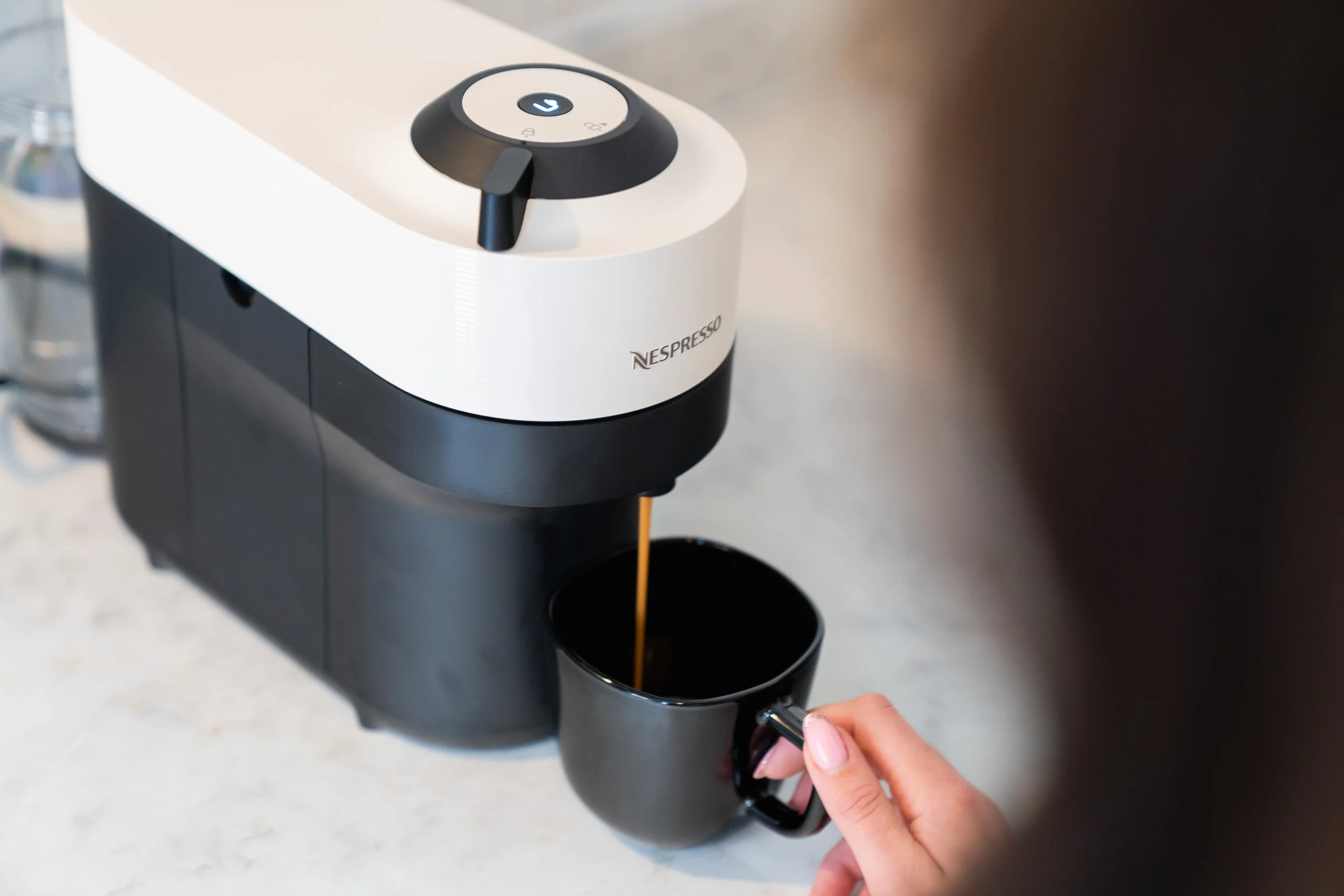 A person holding a black coffee mug under a Nespresso coffee machine, with coffee pouring into the mug.