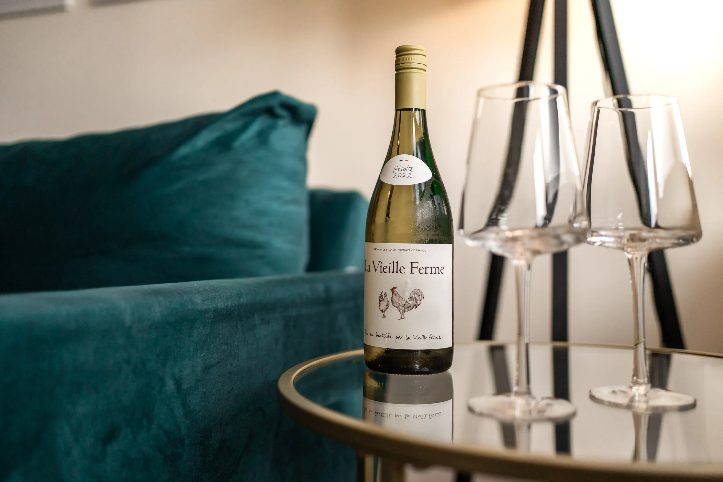 A bottle of La Vieille Ferme wine and three empty wine glasses on a reflective glass table in front of a teal sofa.