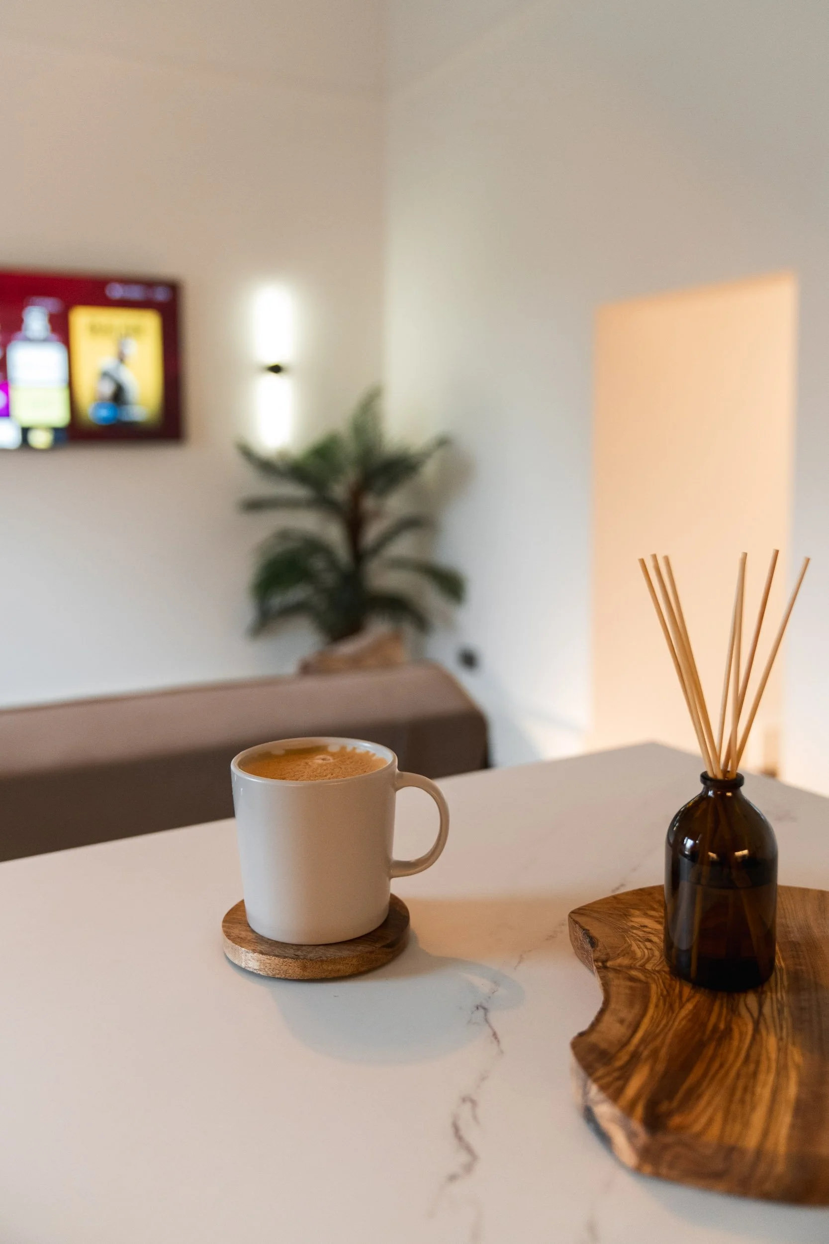A white mug with coffee on a round wooden coaster on a white marble countertop, alongside a dark glass diffuser bottle with reed sticks, on a wooden slab, in a cozy, softly lit room with a TV on the wall and a potted plant in the background.