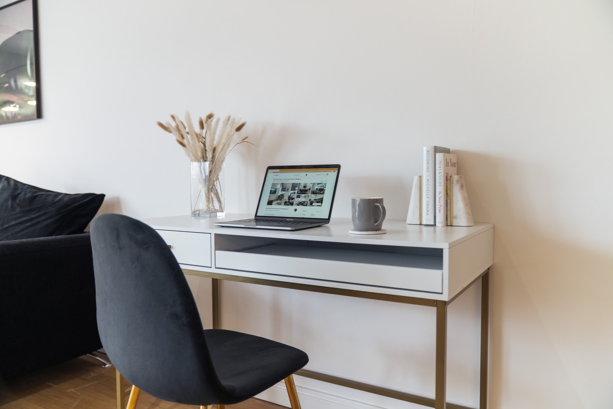 A white desk with a laptop, a gray mug, and a small stack of books in a room with a black chair in front and a decorative vase with dried grass behind.