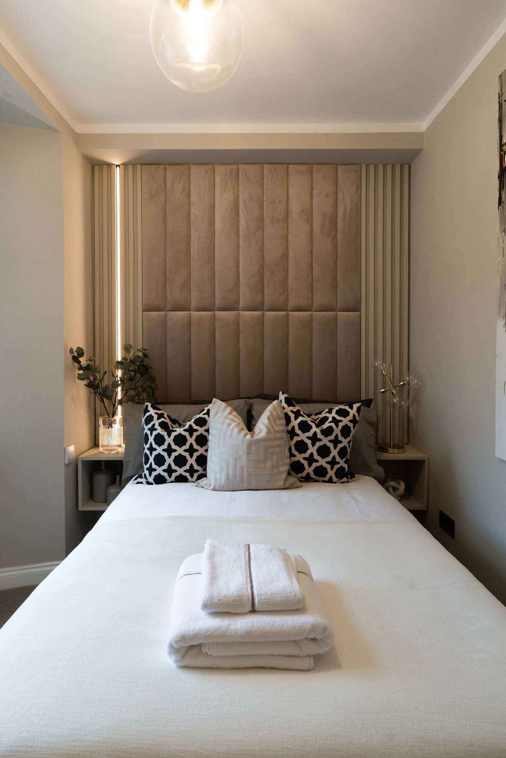 A neatly made bed with white linens, decorated with three patterned throw pillows in black, white, and beige, with folded towels on top, and an upholstered headboard with beige vertical panels. There are two bedside tables with decorative items inclu