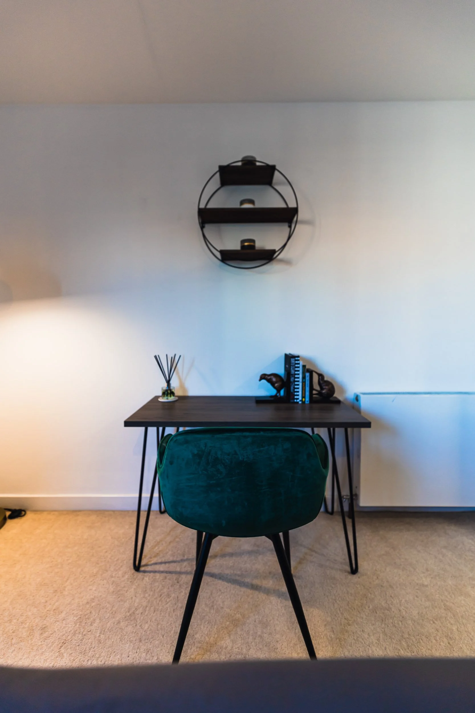 A modern desk with a green velvet chair, decorative objects, books, and a floating wall shelf on a white wall.