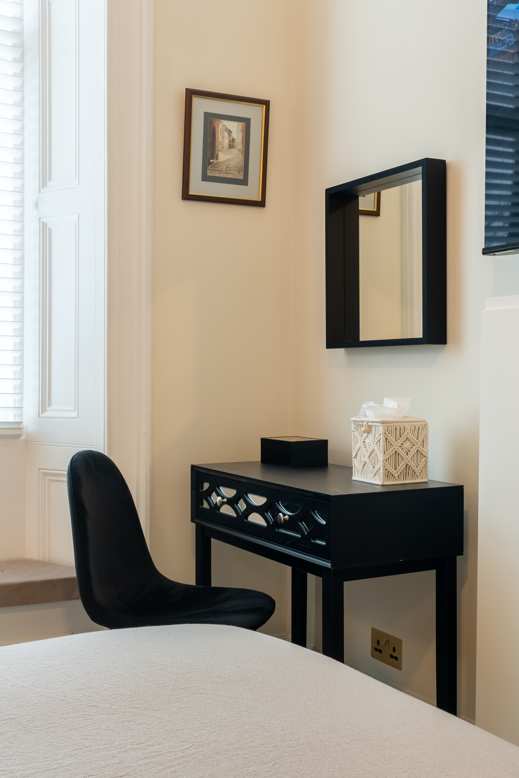 A small black table with tissue box and decorative box on top, black mirror on the wall above, black chair to the left, framed artwork on the beige wall, window with white shutters on the left, and part of a bed in the foreground.