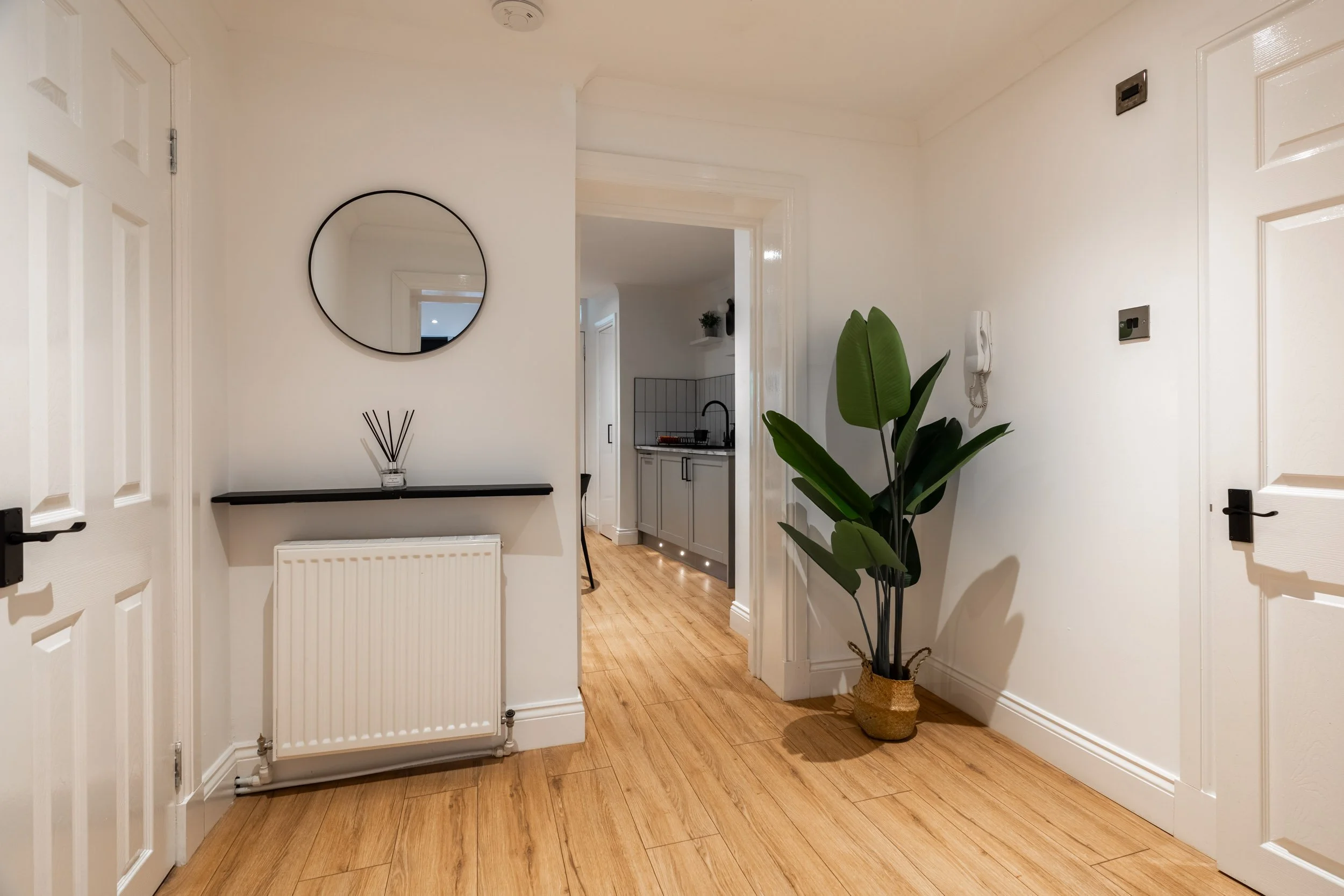 Interior view of a hallway leading to a kitchen, with a potted plant, wall mirror, and a shelf against white walls and wooden flooring.