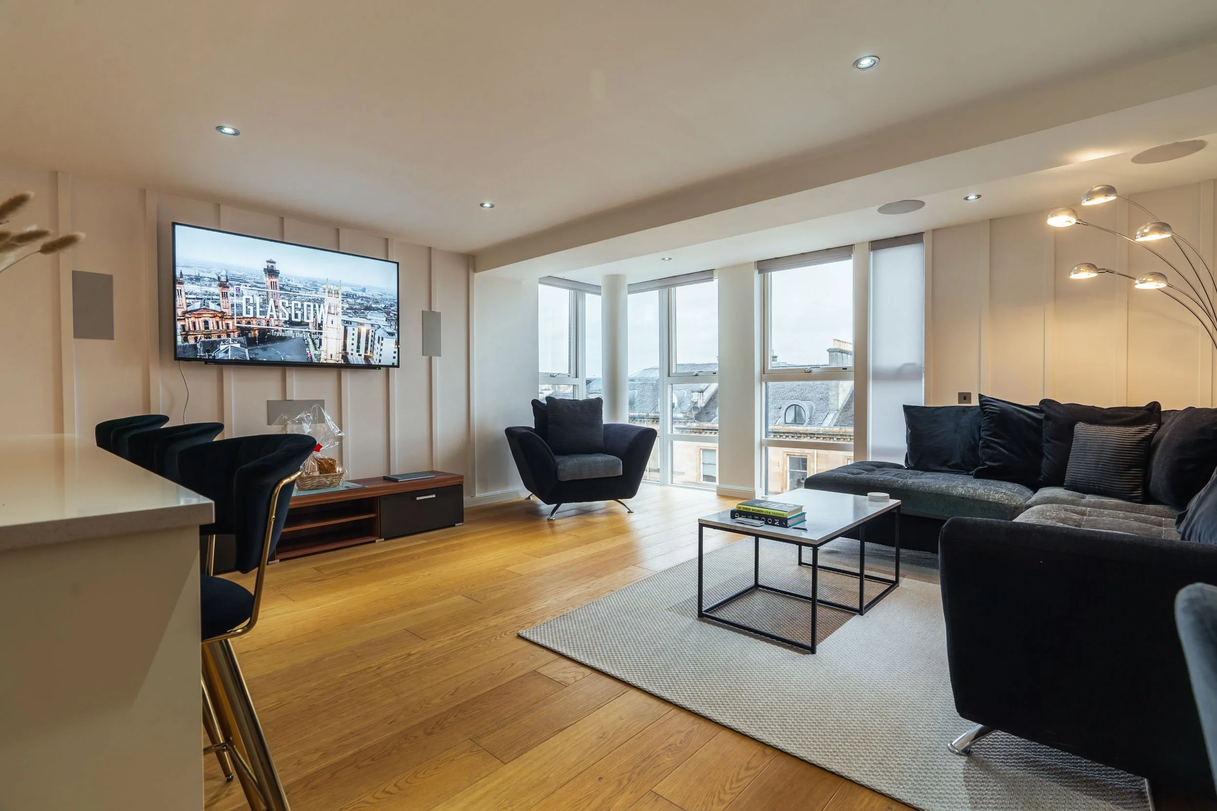 Living room with large windows, black sectional sofa, black armchair, glass coffee table, TV on wall displaying 'Glasgow', and a floor lamp with multiple bulbs, wooden floor, and a beige area rug.