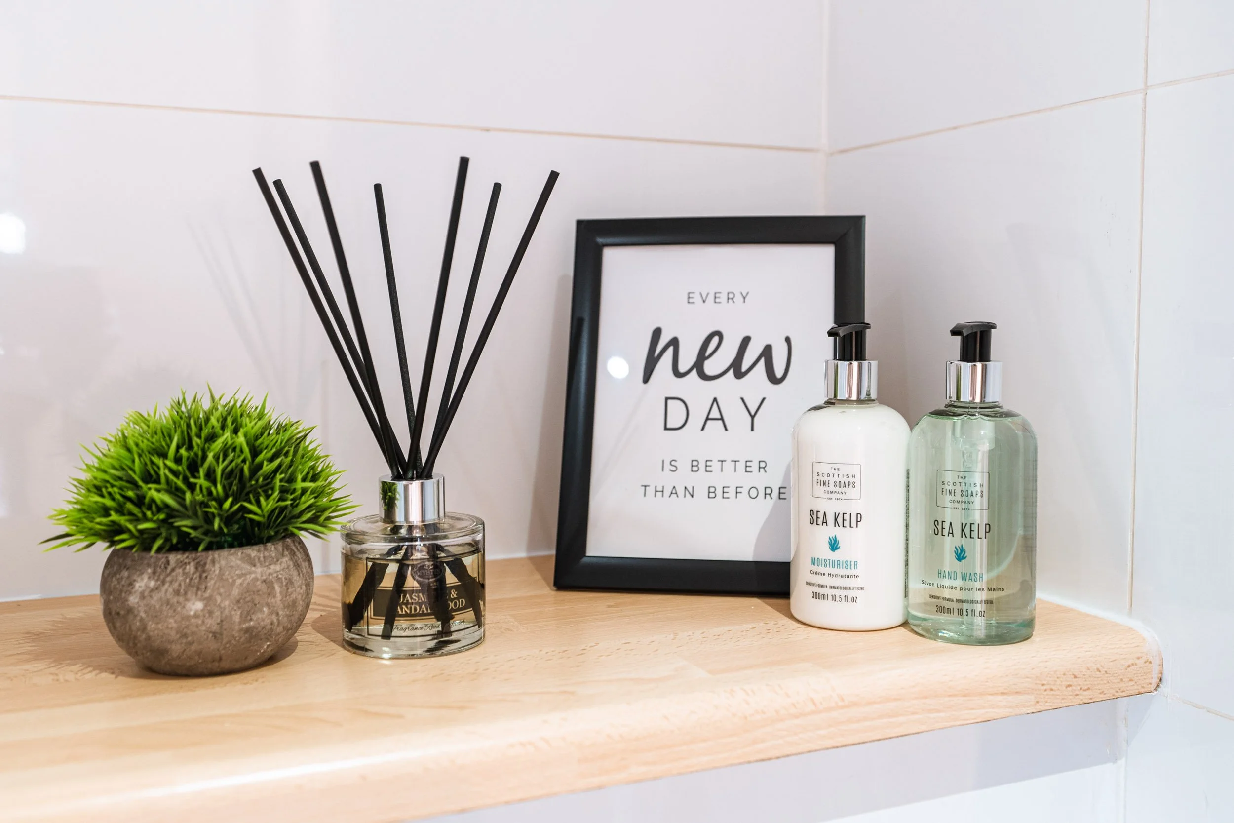 A bathroom shelf with a potted green plant, a reed diffuser, a framed quote, and two bottles of Sea Kelp hand soap and moisturizer.
