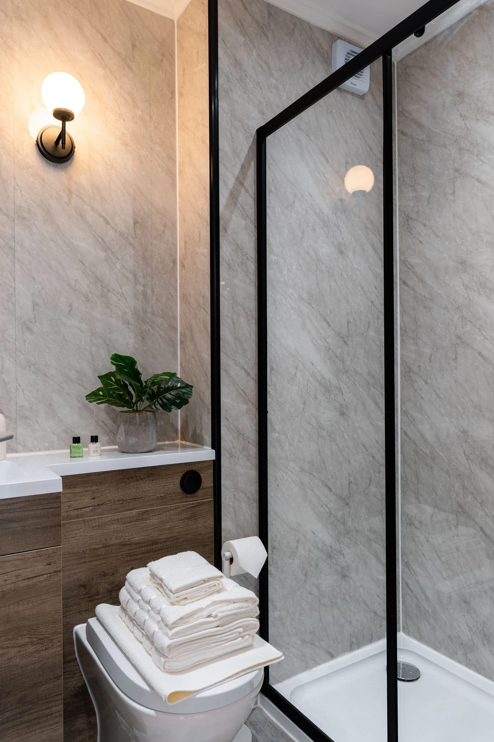 A modern bathroom with a glass shower enclosure, a wall-mounted light fixture, a potted plant on a wooden vanity, and a stack of neatly folded towels on top of the toilet.