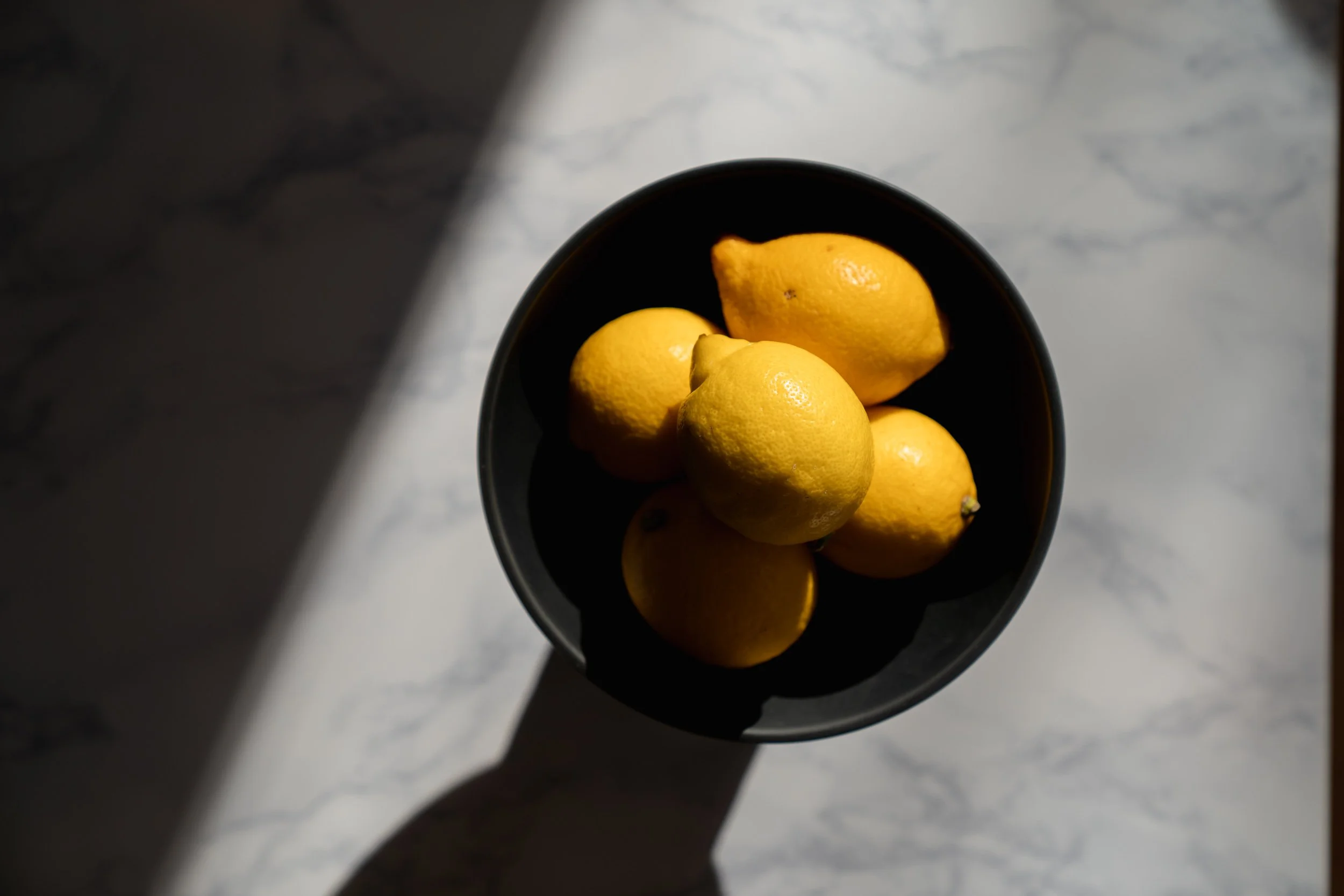 A black bowl containing six lemons on a white marble surface, with shadow and sunlight highlighting the lemons.