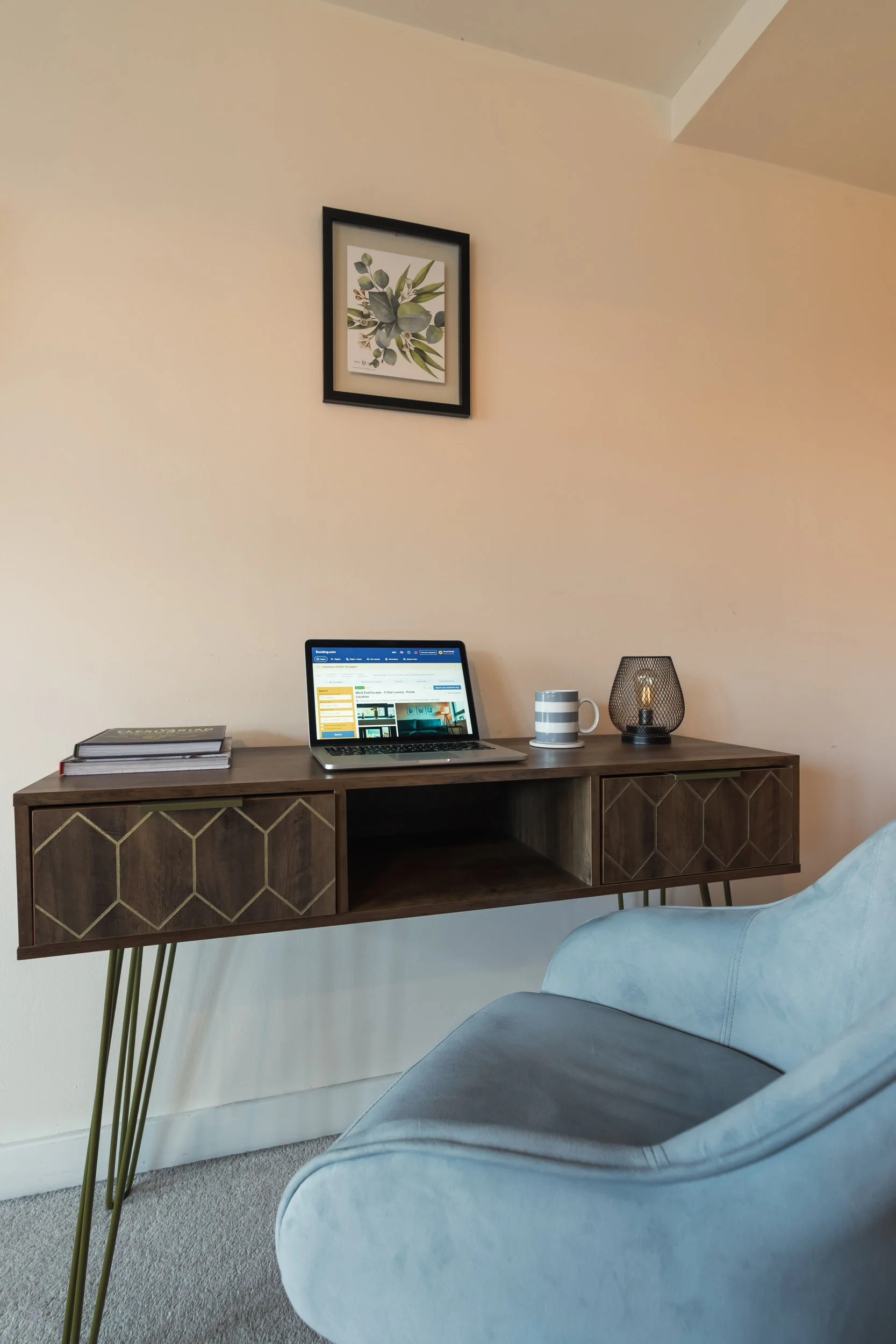 A wooden desk with gold geometric accents on the cabinet doors, holding a closed book, an open laptop displaying a website, a striped mug, and a black wire lamp with a visible filament bulb. A light blue armchair is visible in the foreground. A frame