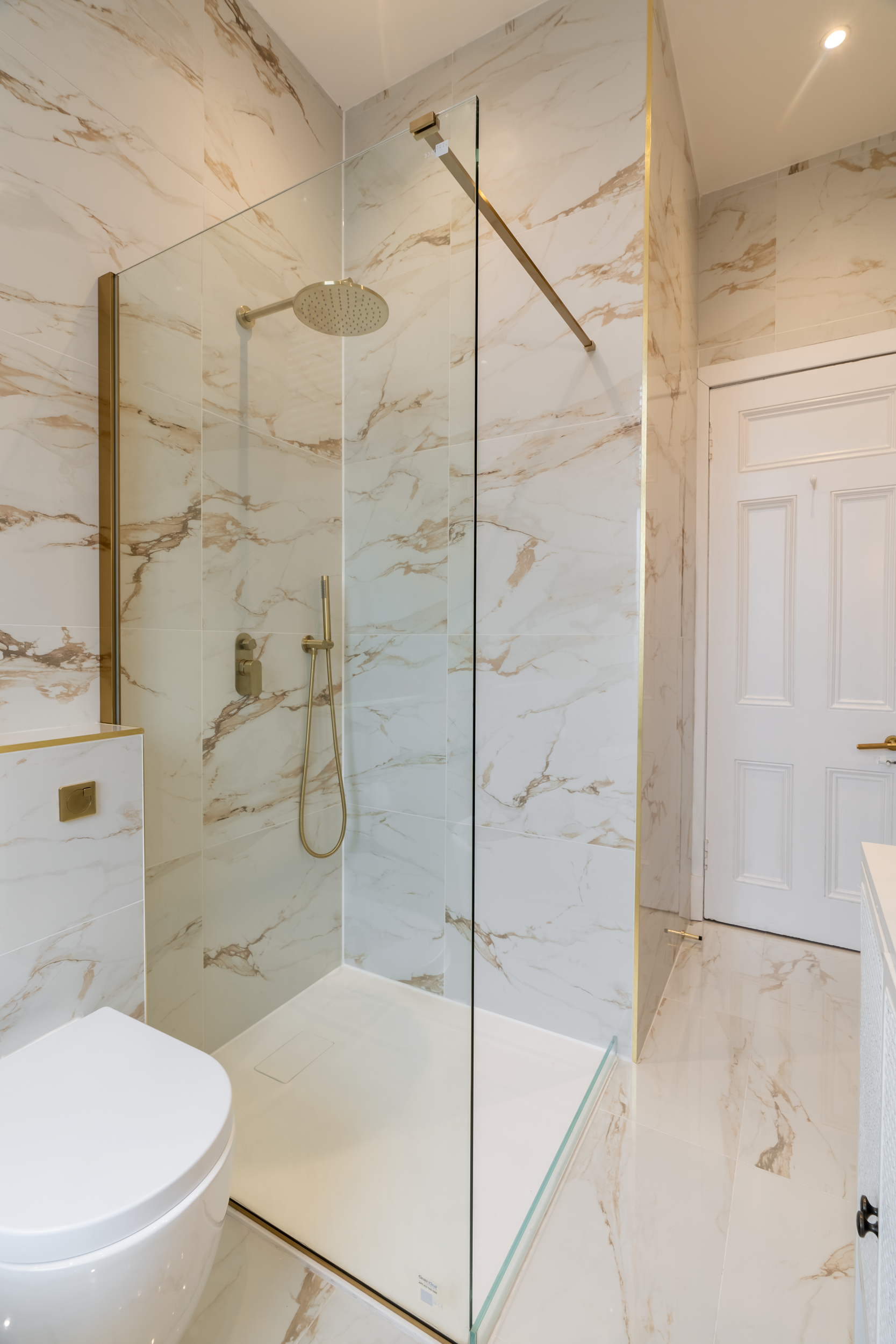Modern bathroom with marble walls and floors, glass shower enclosure, rain showerhead, handheld shower, white toilet, and white door.