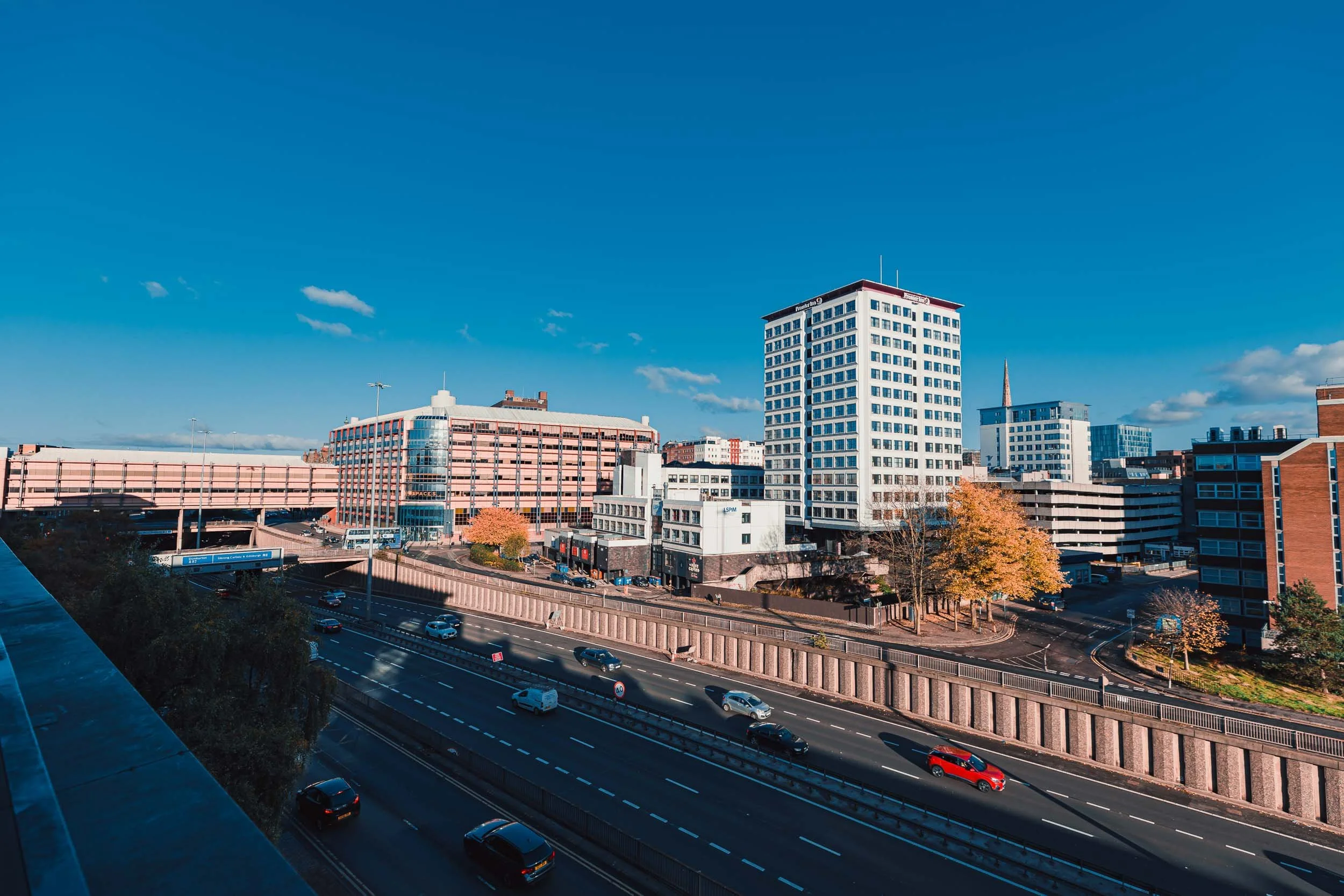 Cityscape with multiple buildings, including a tall white building, a parking structure, and a highway with cars, under a clear blue sky.