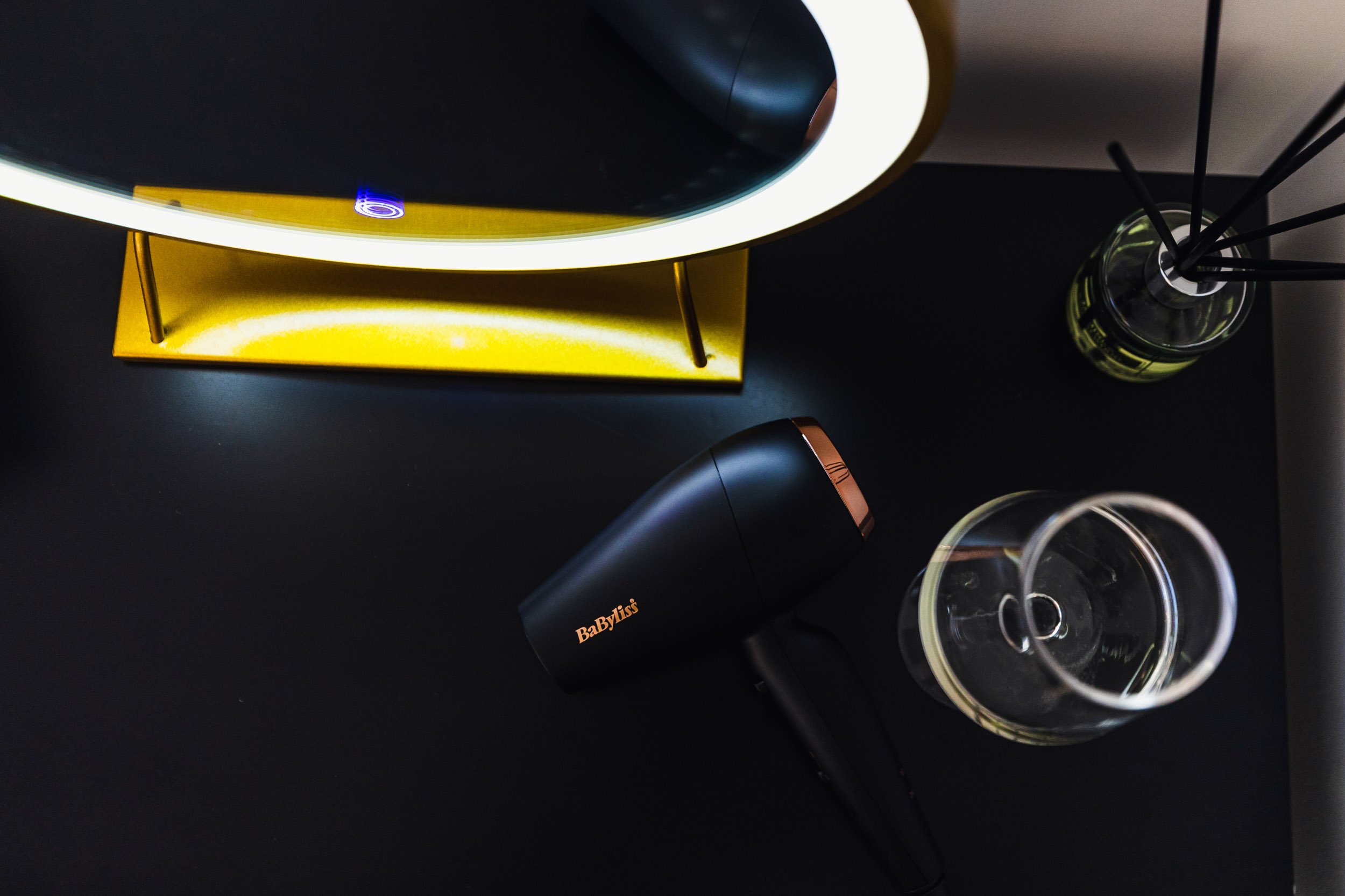 A top-down view of a black table with a yellow lamp, a black BaByliss hairdryer, two empty wine glasses, and a reed diffuser with black sticks.