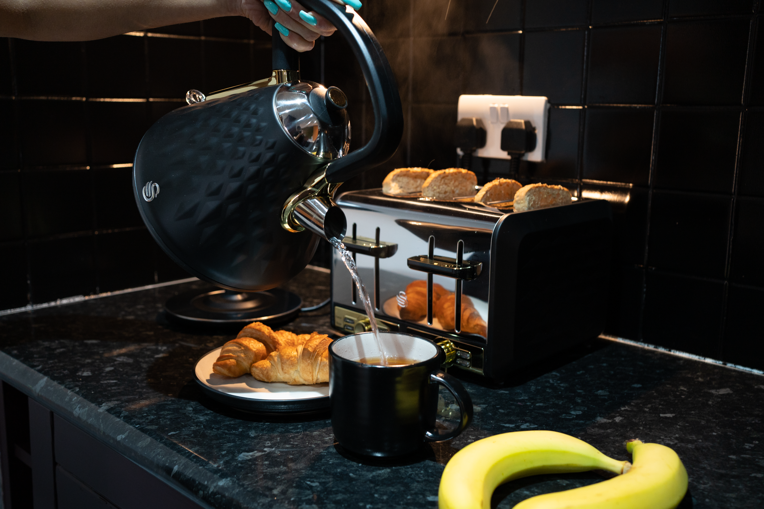 Black and gold electric kettle pouring hot water into a black mug on a kitchen countertop. A plate with croissants and a banana nearby. A toaster with slices of bread and croissants on it. Black tiled wall in the background.