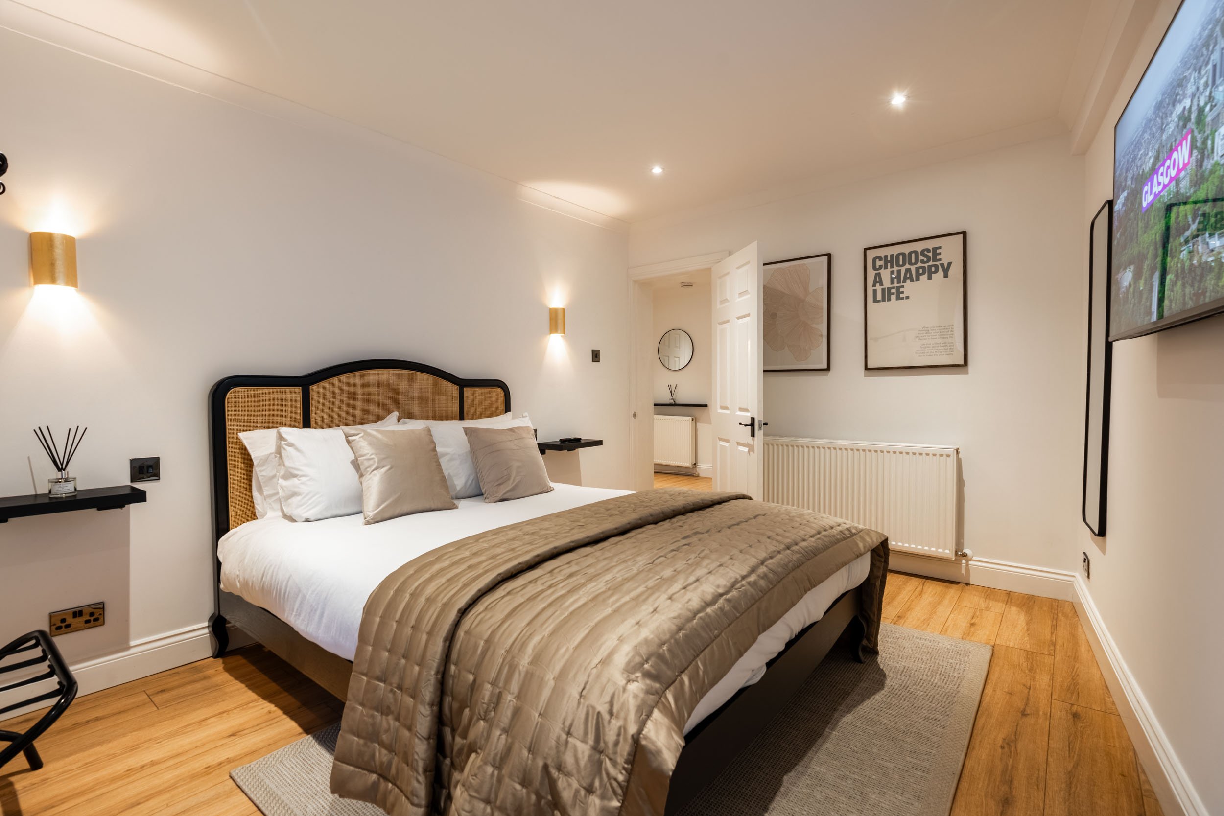 A neatly made bedroom with a bed that has a black frame and a woven headboard, white bedding, and beige pillows. The room has white walls, wood flooring, and minimal decor, including framed wall art and a mirror. A flat-screen TV is mounted on the wa