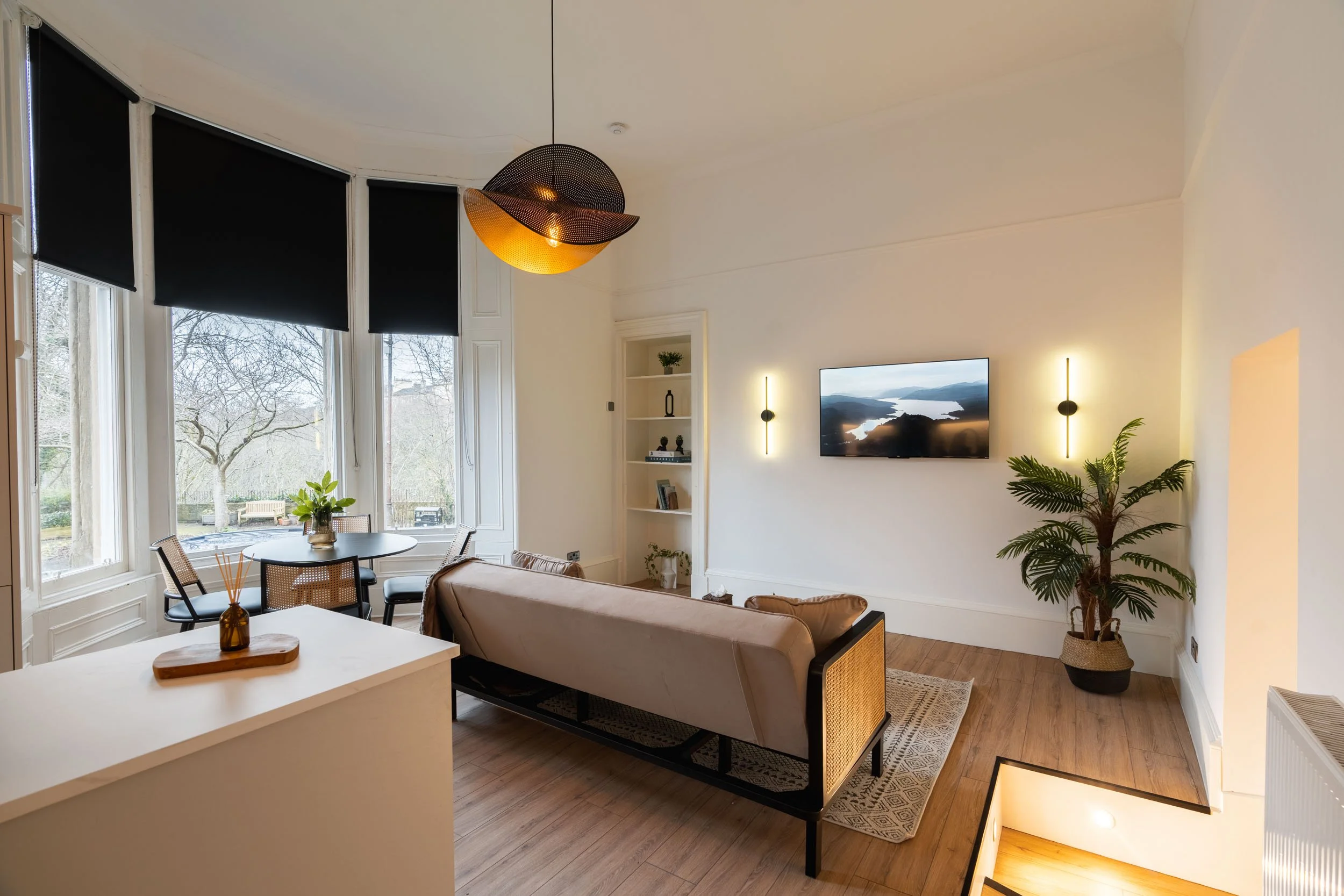 Bright living room with large windows, black shades, and a sofa facing a wall-mounted TV. There is a round dining table with chairs near the windows, a potted plant, built-in shelves, and modern wall sconces providing accent lighting.