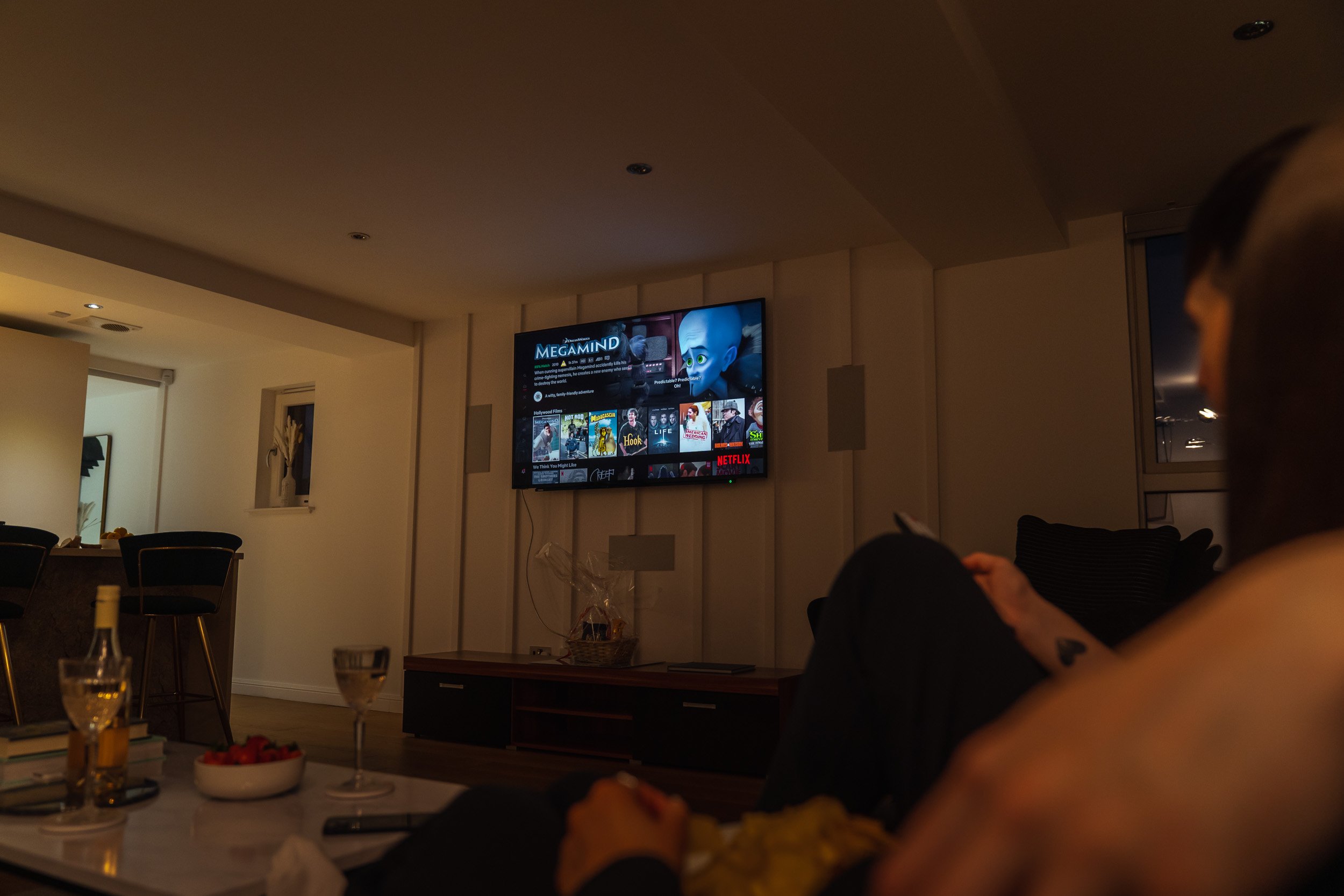Living room with a television showing the Netflix homepage, two people sitting on a sofa, a table with glasses and snacks, and a kitchen in the background.