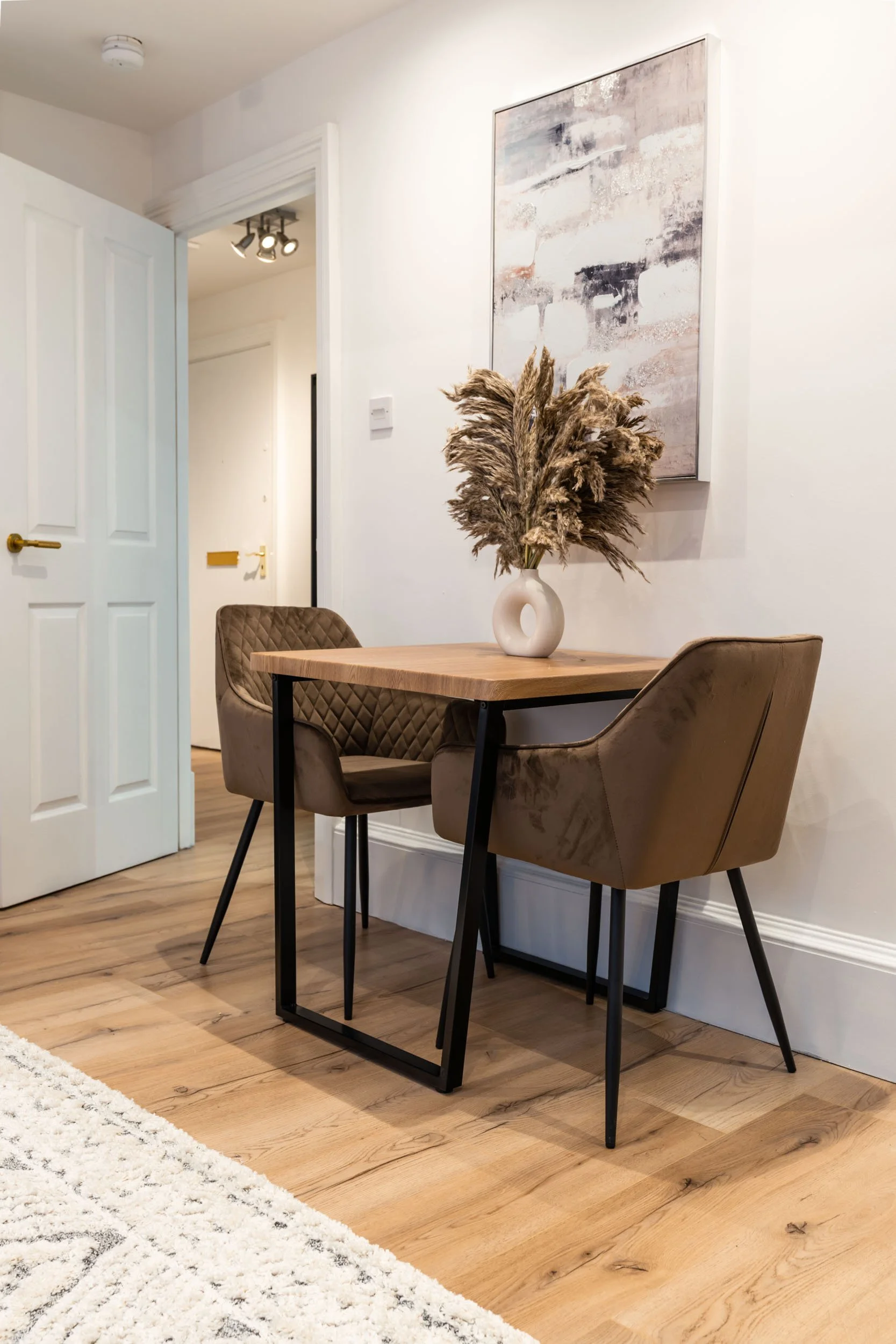 A small wooden dining table with a white vase containing dried pampas grass, two beige chairs, and a large abstract art piece on a white wall in a well-lit room with wood flooring.