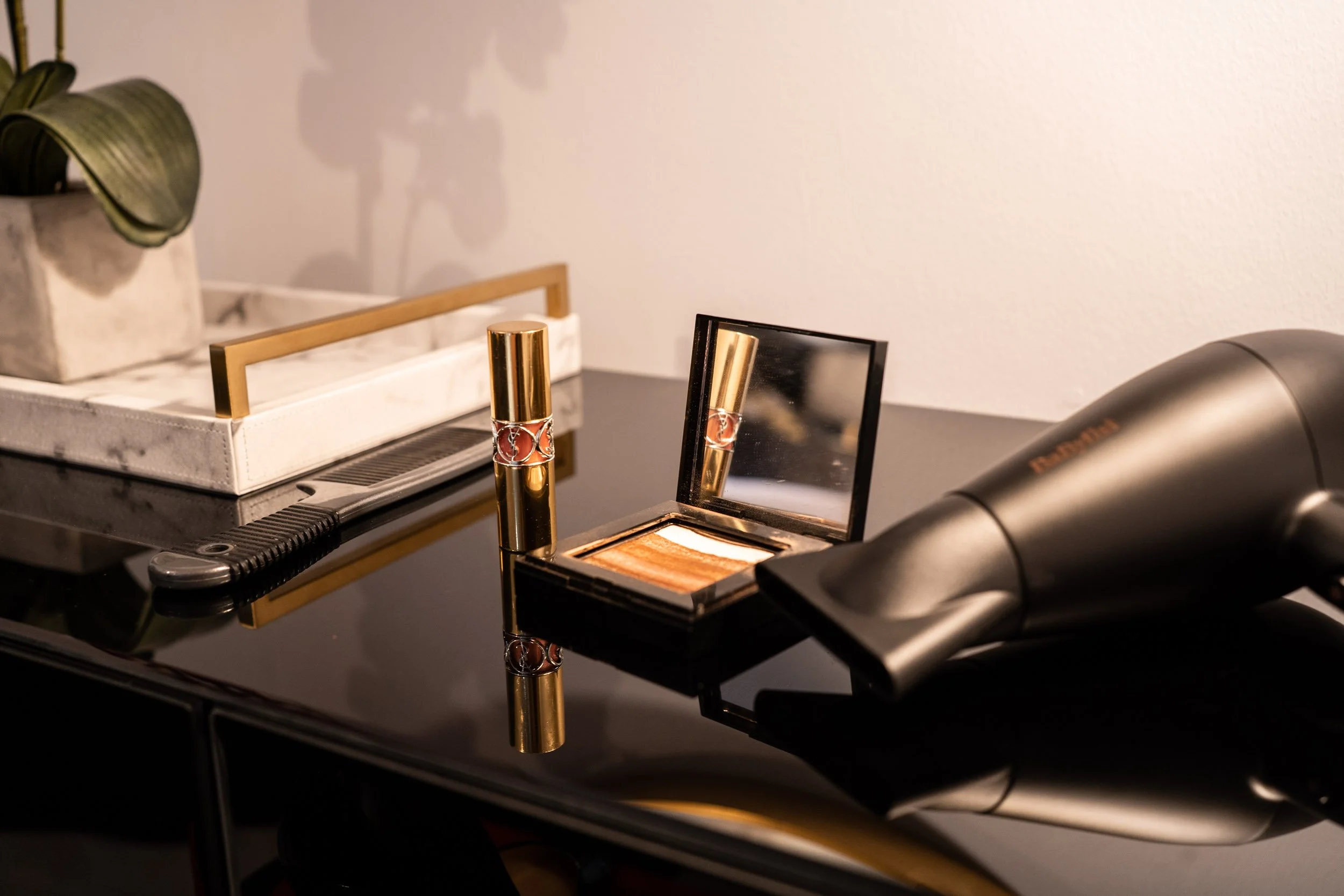 Beauty products on a black table, including a lipstick, eyeshadow palette, hair dryer, and a decorative plant on a tray.