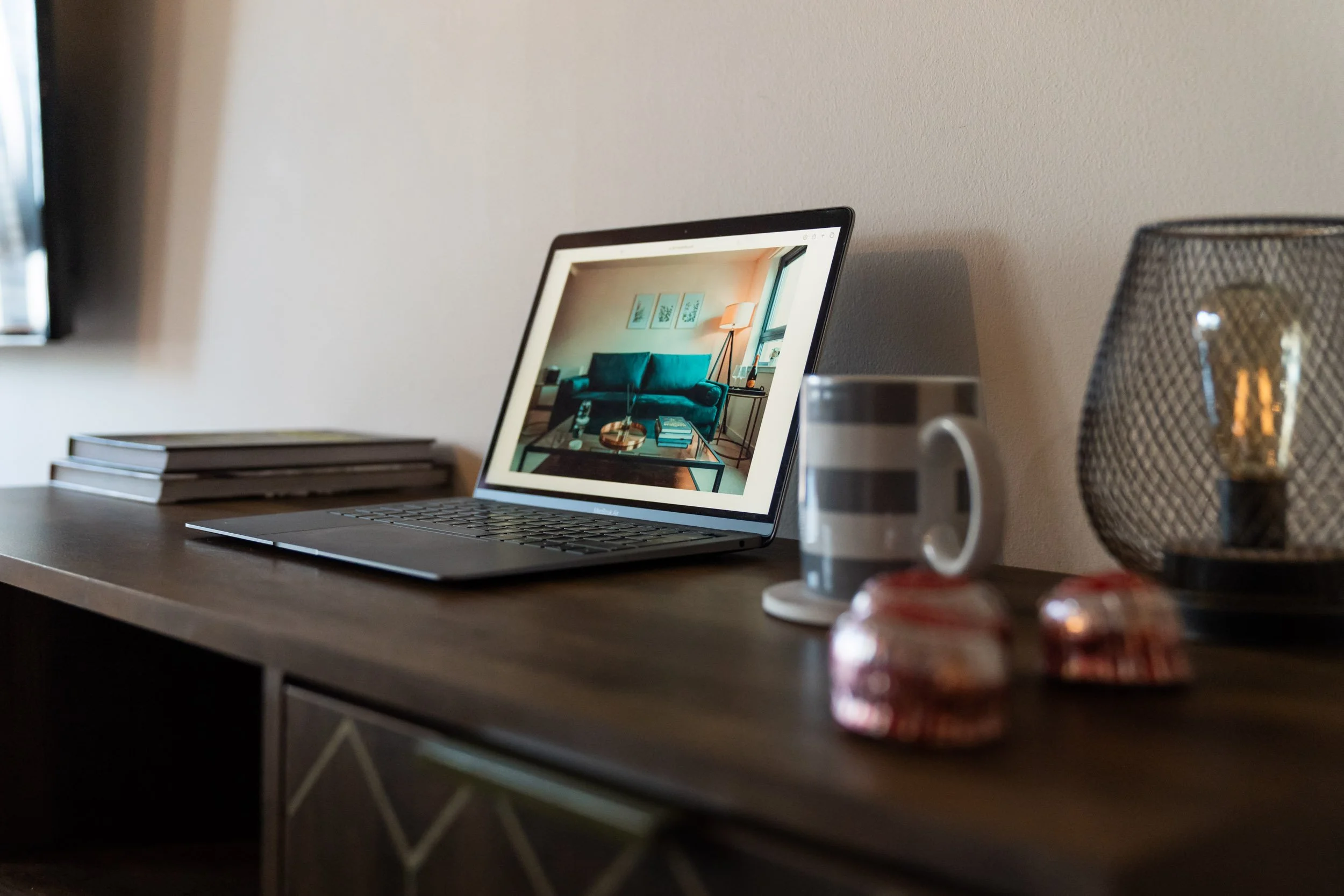 A laptop on a wooden desk displaying a photo of a living room with a teal sofa, side table, and framed artworks. Next to the laptop are a striped mug and a glass jar with a light bulb, with extra stacked notebooks on the left.