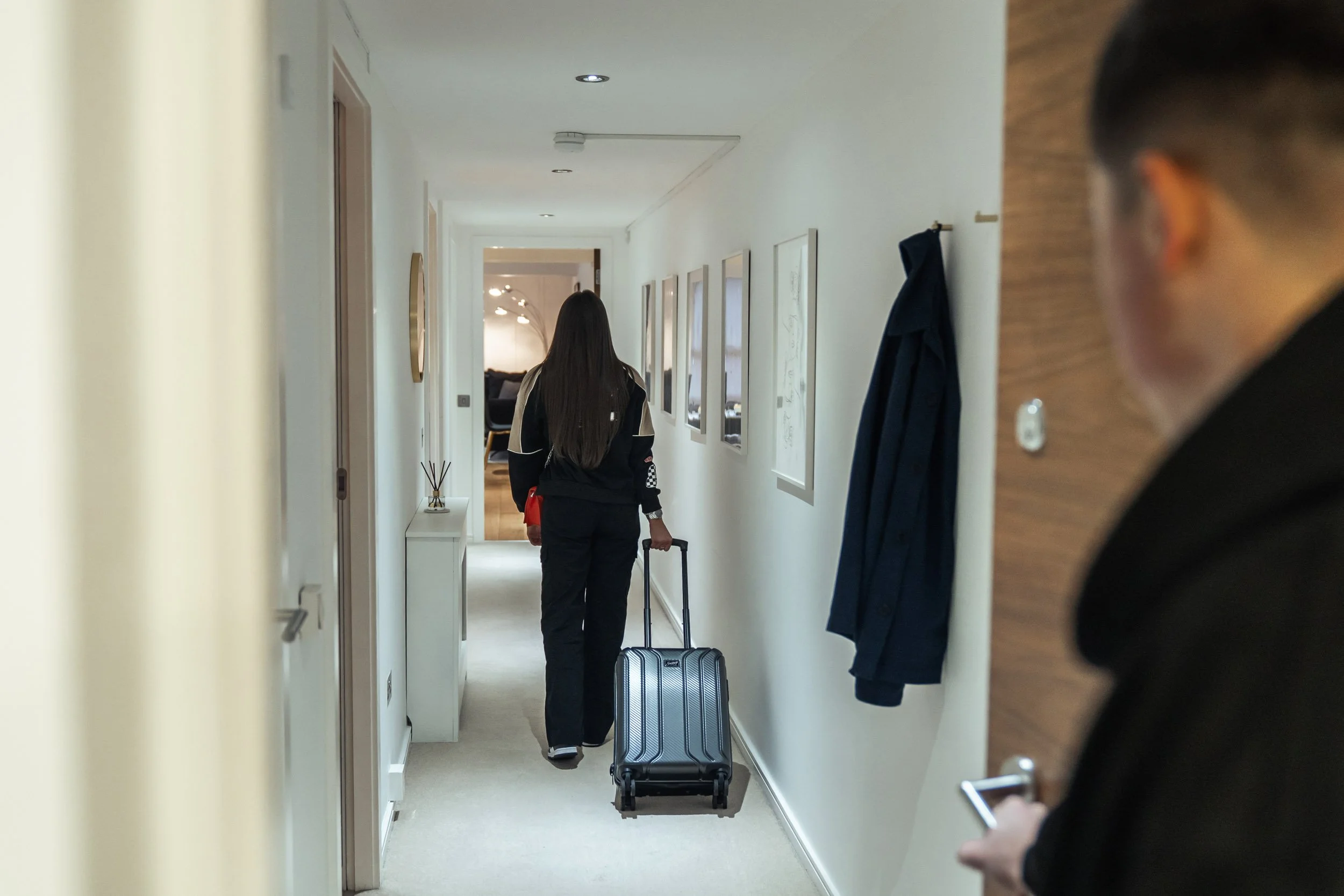 A woman walking down a hallway pulling a suitcase, with a man at the door holding a phone.