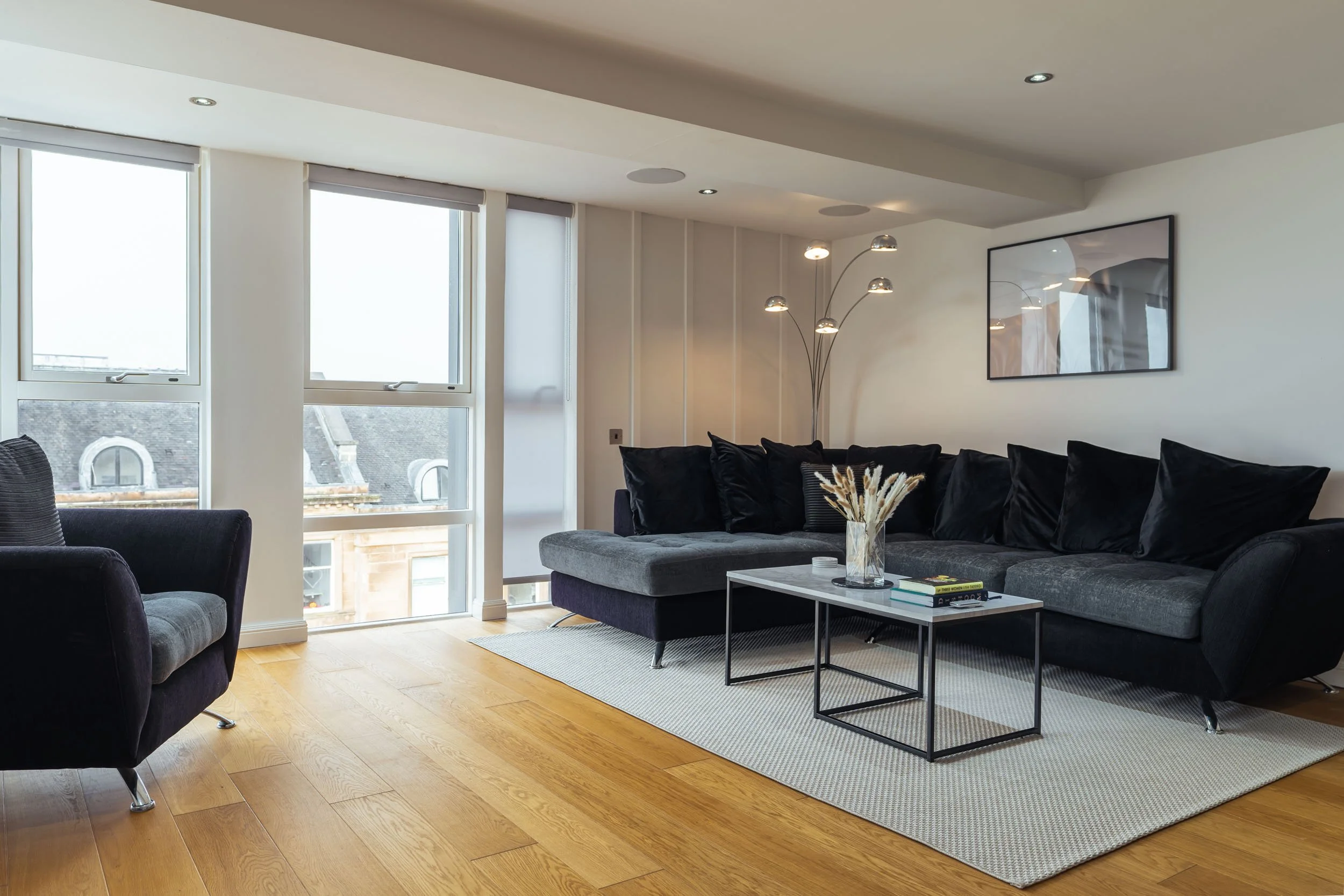 Modern living room with black and gray sectional sofa, black armchair, wooden floor, white walls, large windows, abstract wall art, and a black and white area rug.
