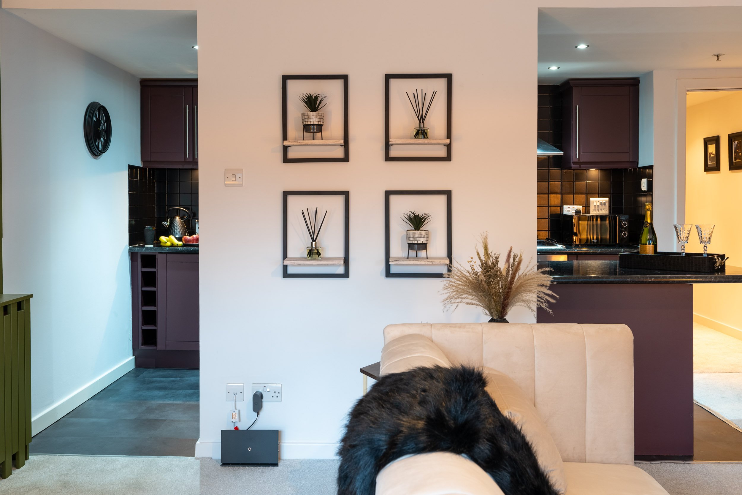 Living room with a beige sofa, wall decoration of four framed pictures with plants and reed diffusers, a vase with dried flowers on the side table, and a partially visible kitchen with dark cabinets and black backsplash.