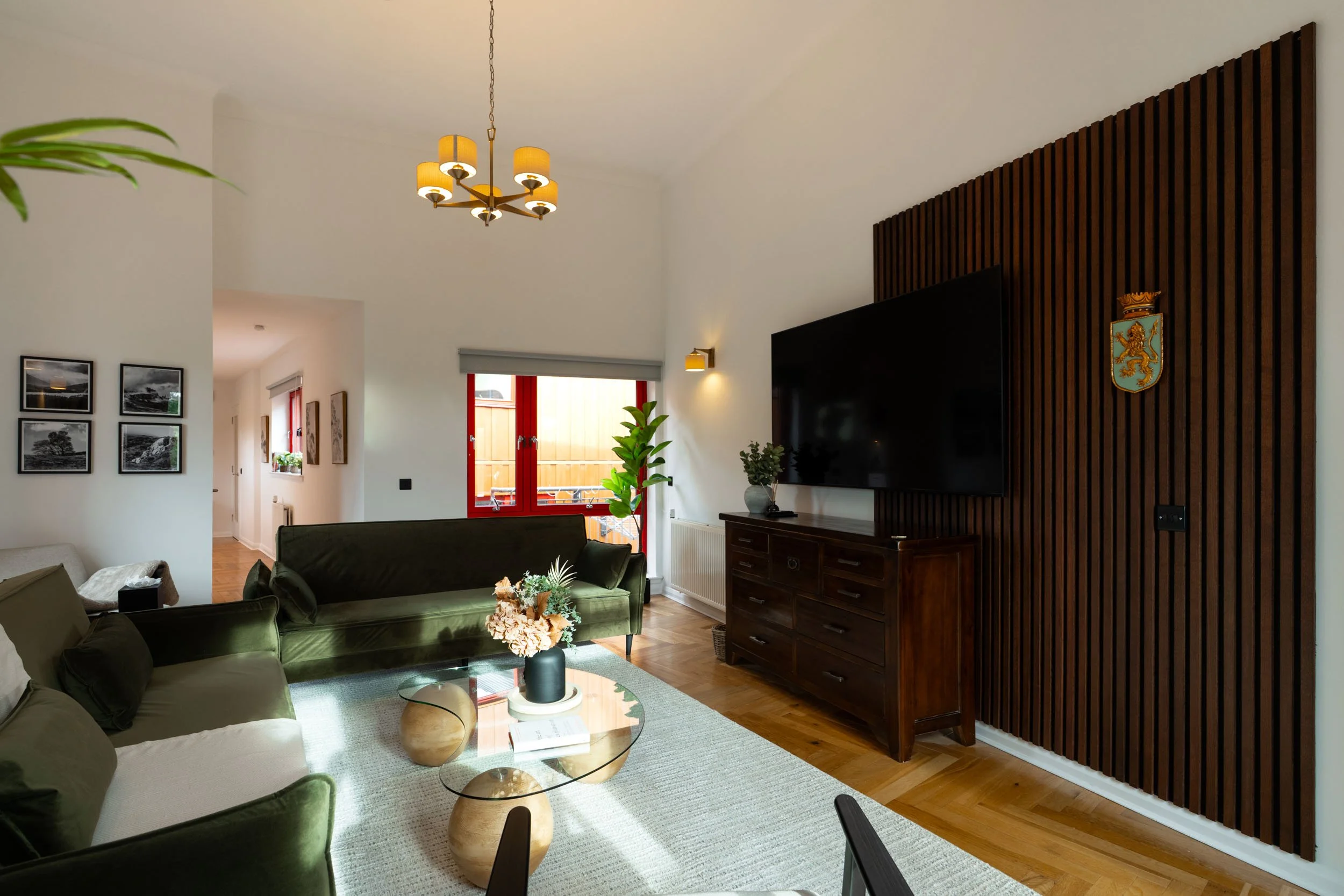 Living room with white walls, green velvet sofas, a glass coffee table, a wooden sideboard with a TV, and a decorative wooden panel with a crest, during daytime.