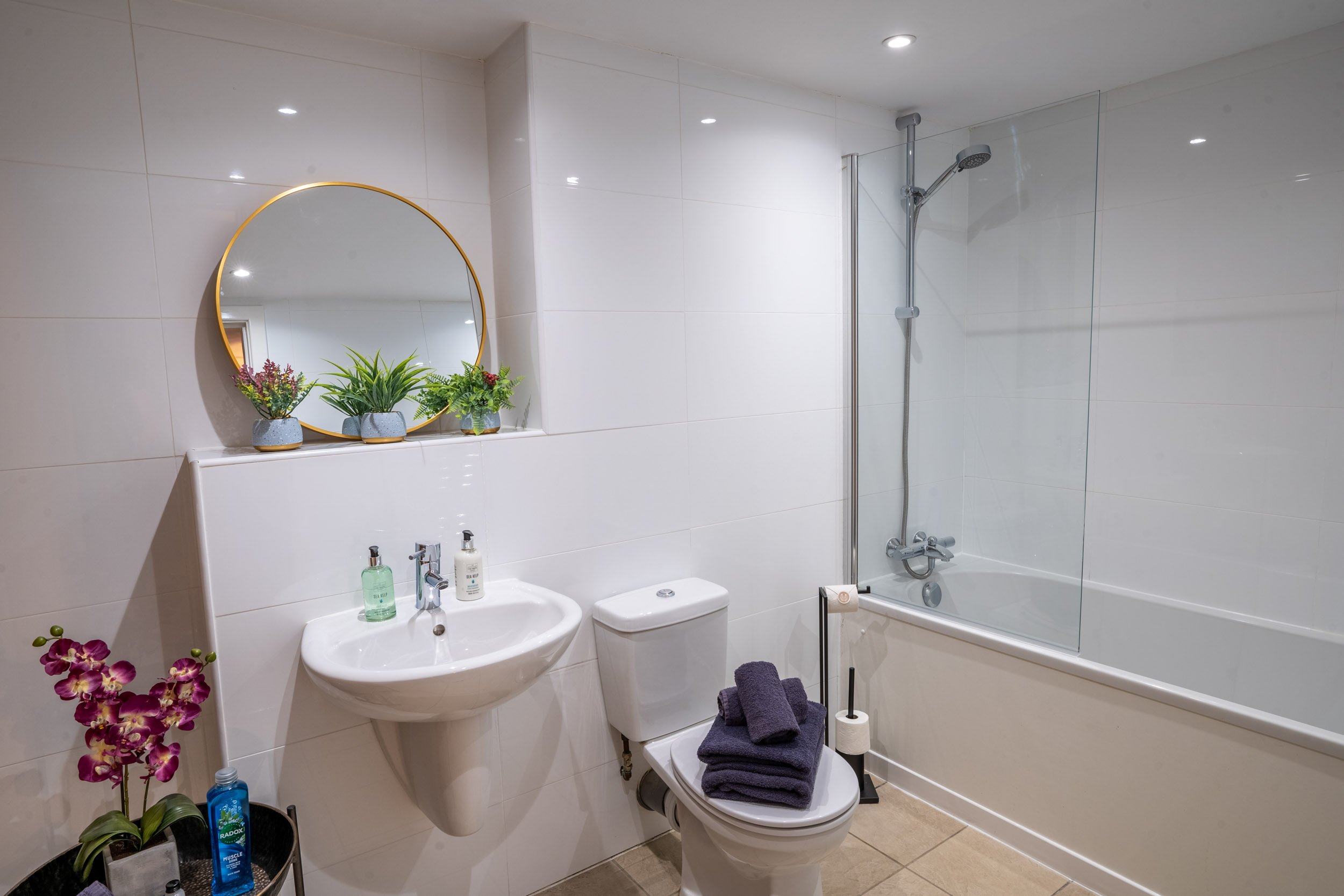 Bathroom with white tiled walls, a round mirror above a small sink with soap dispensers, a toilet with folded towels, a plant shelf, a pink orchid, and a bathtub with a glass shower screen.