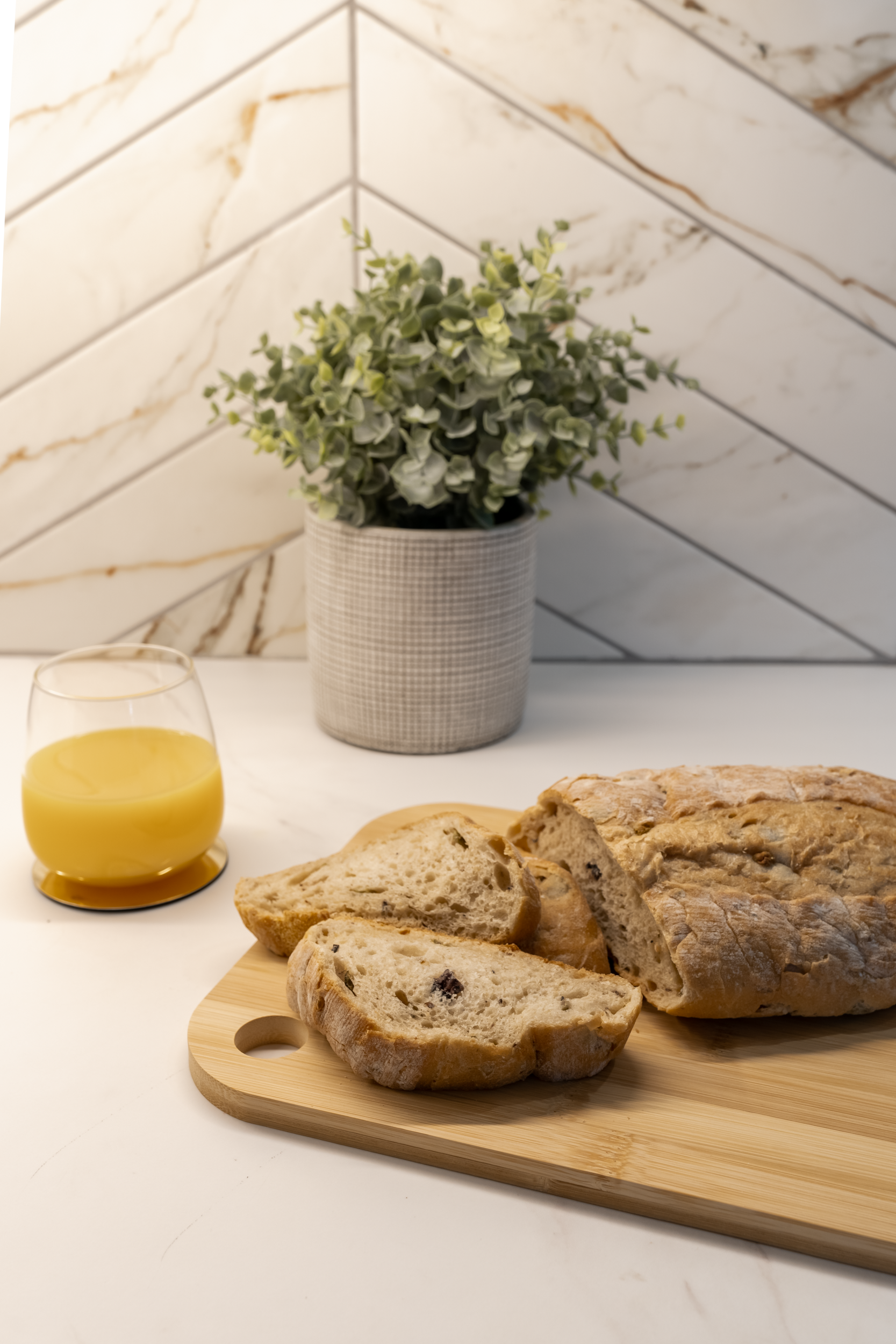 A loaf of bread with slices cut on a wooden cutting board next to a glass of orange juice, with a green potted plant and a tiled wall in the background.