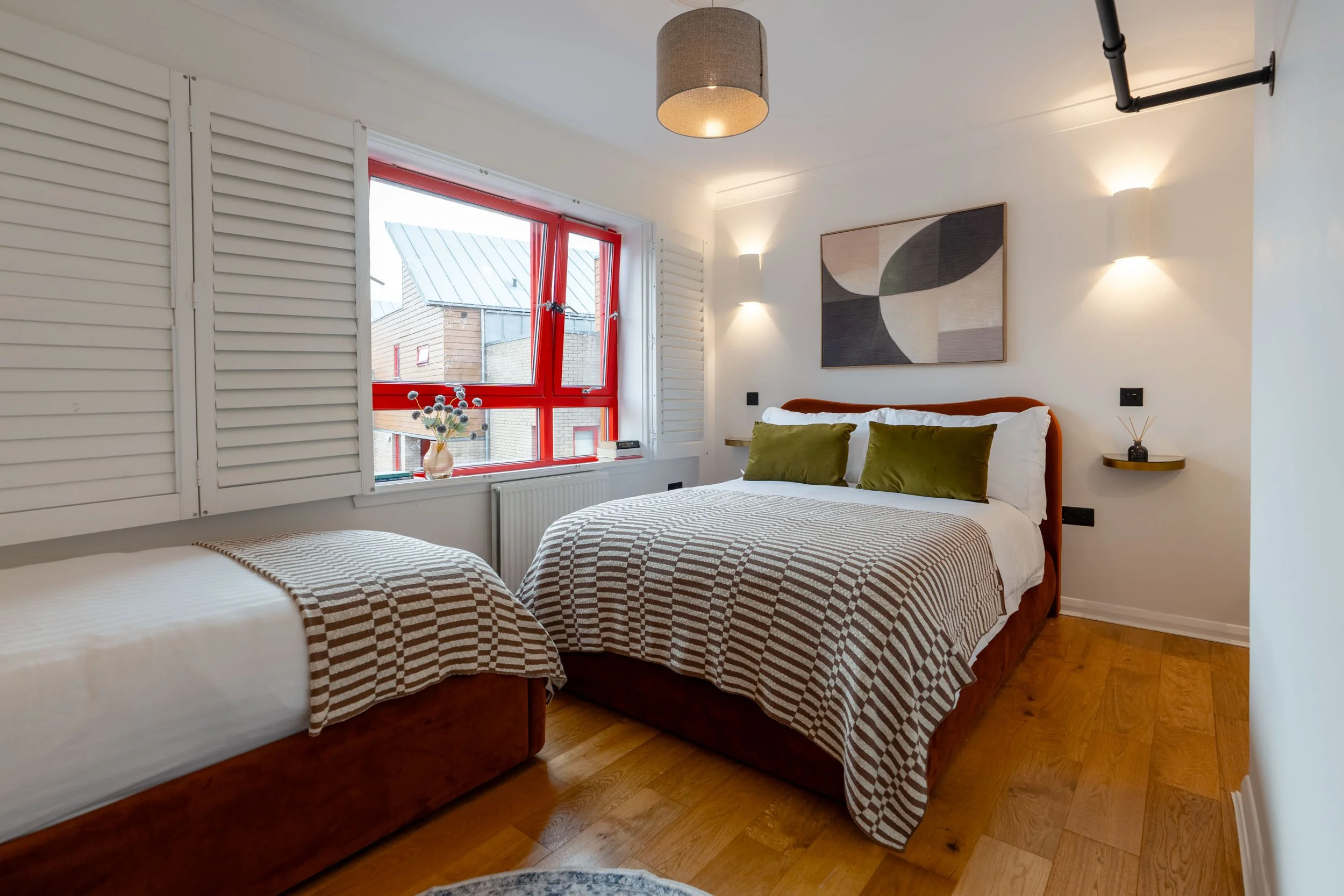 A bedroom with a large bed, a smaller bed, wooden floors, a window with red frame, white shutters, and minimal decor.