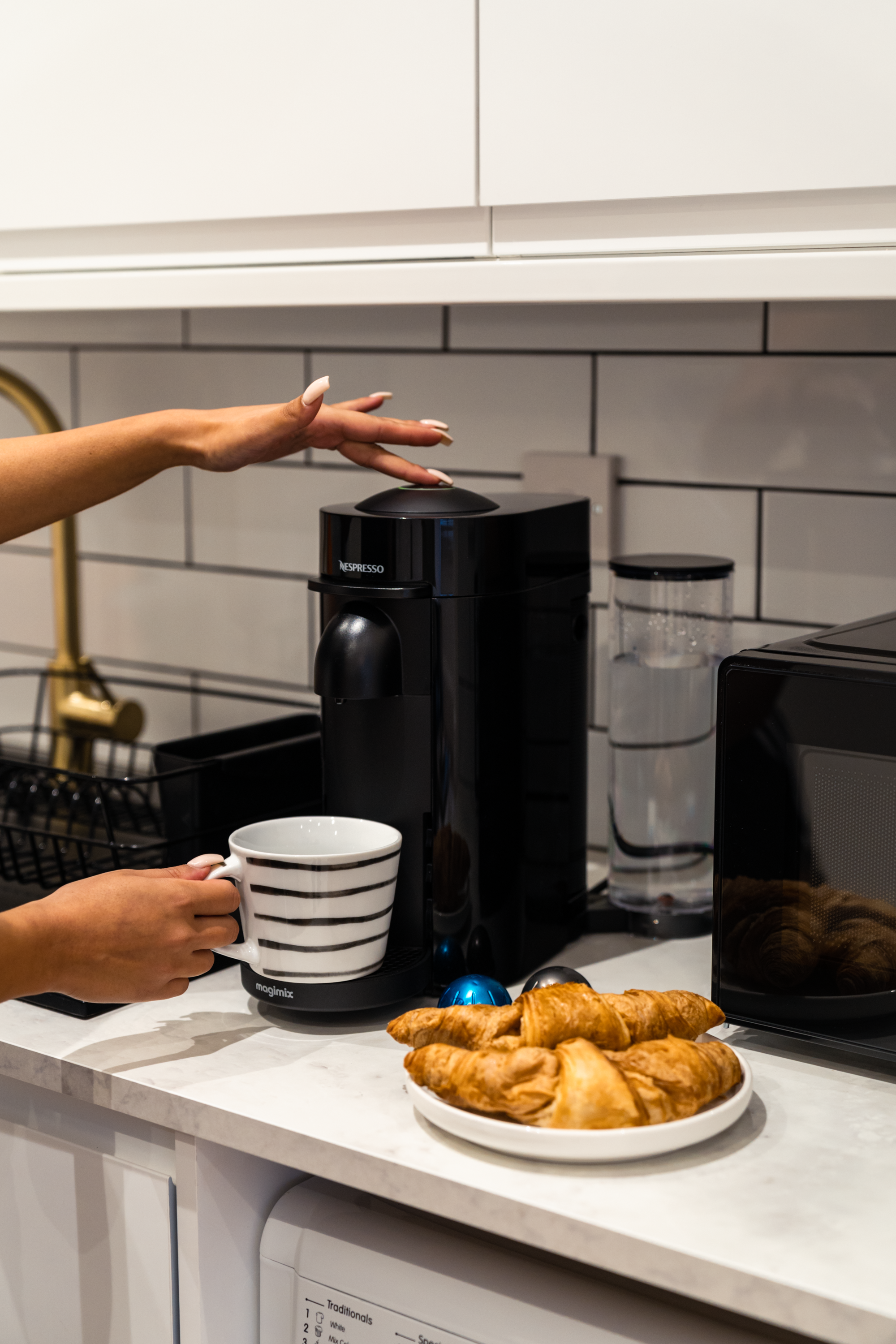 A kitchen countertop with a Nespresso coffee machine, a striped mug, a plate of croissants, a water pitcher, and a black microwave oven.