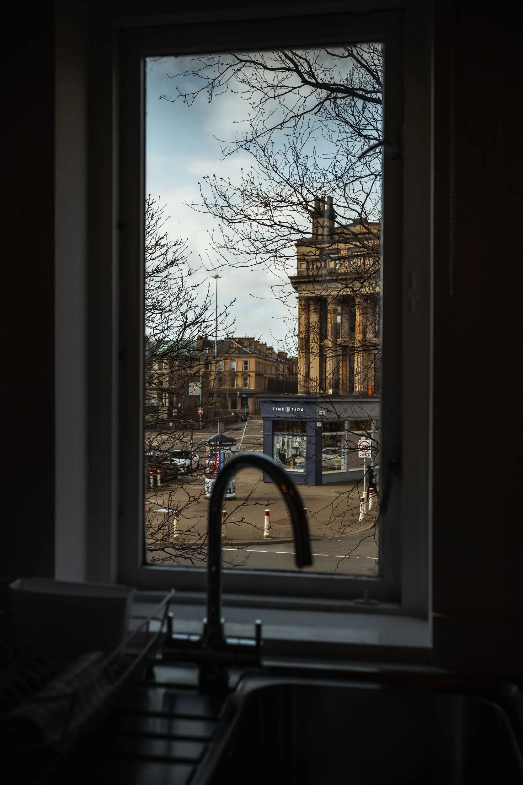 View through a kitchen window showing a city street with trees, buildings, and a storefront named 'Time @ Tine'.
