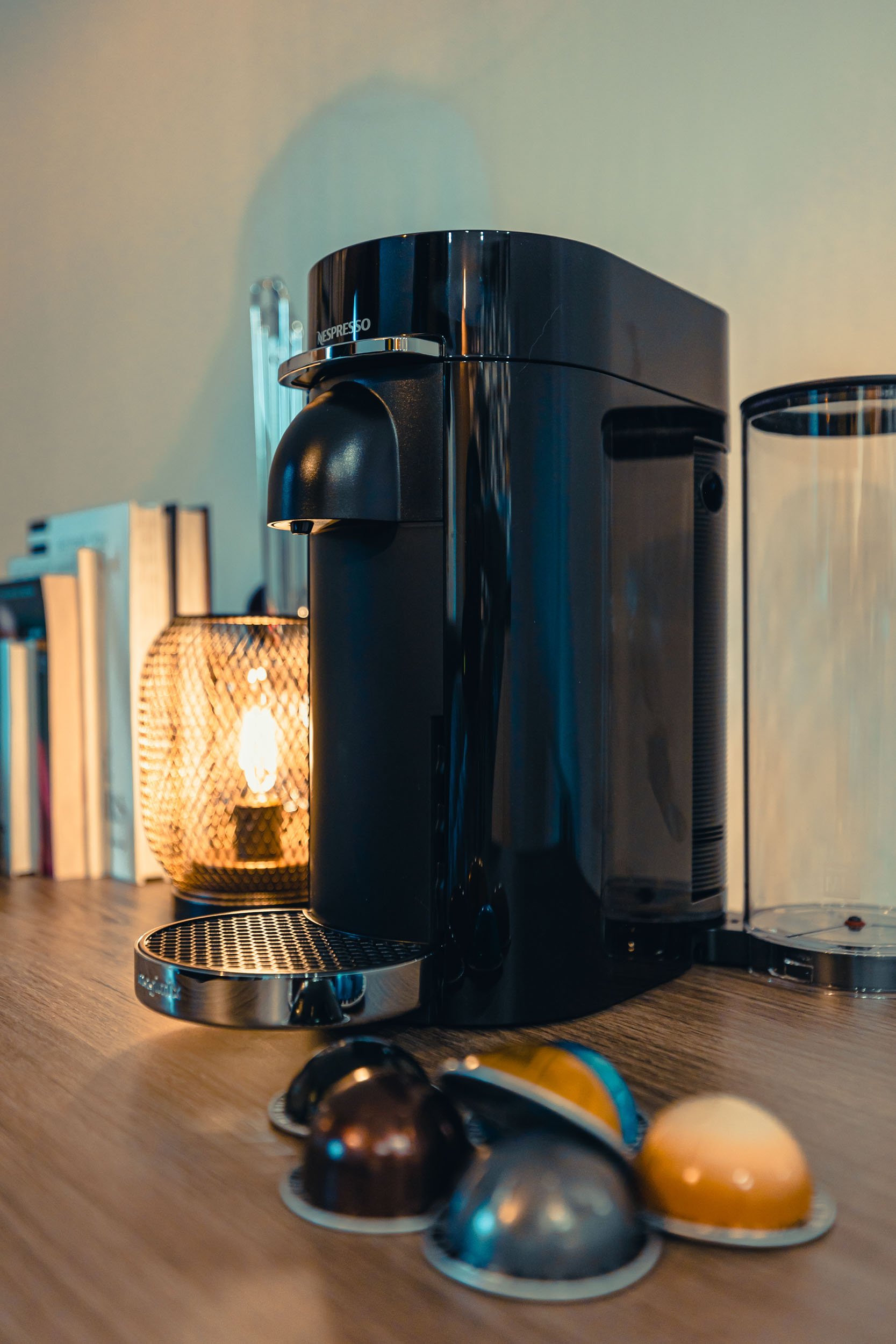 Black Nespresso coffee machine on a wooden countertop with coffee capsules in front, a lit candle in a wire holder, and books on the side.