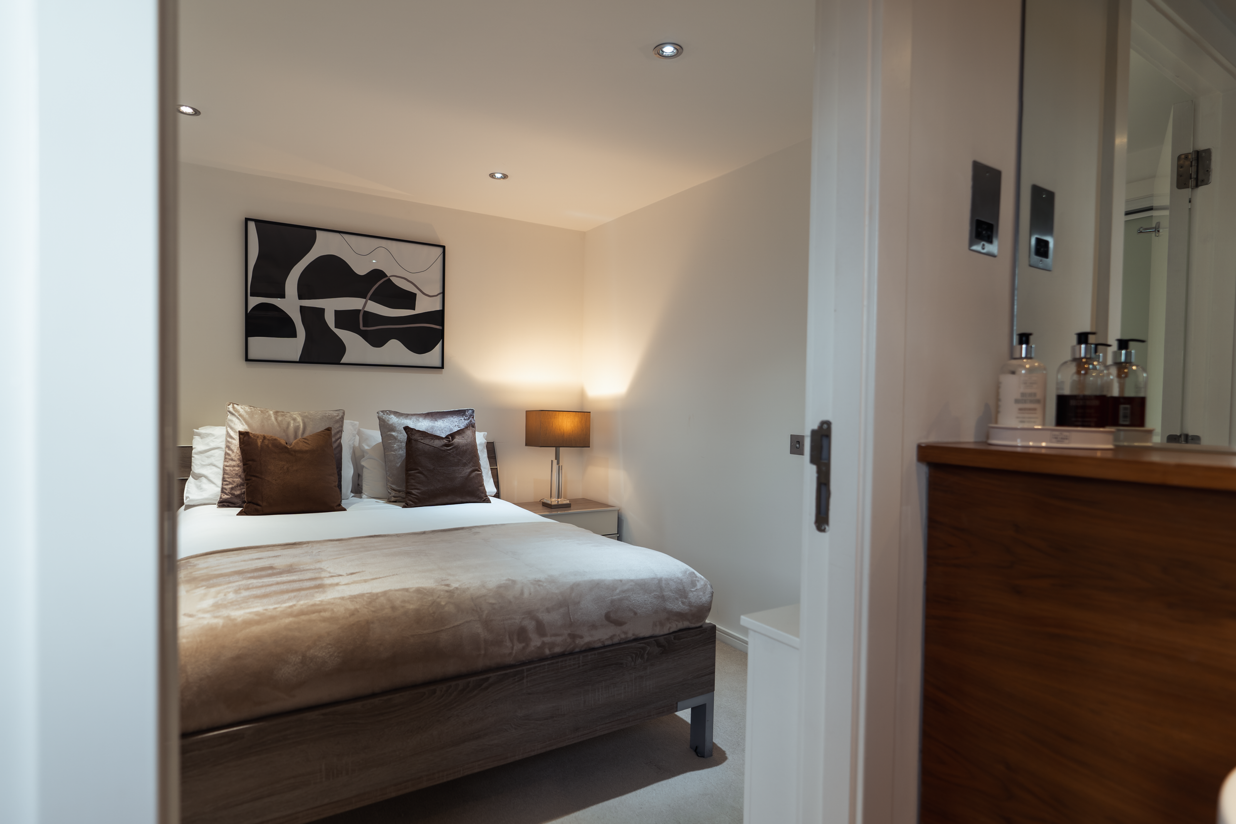 A neatly made bed with multiple pillows against a beige wall, a black and white abstract art piece above, a bedside table with a lamp, and a wooden dresser with bottles of toiletries nearby.