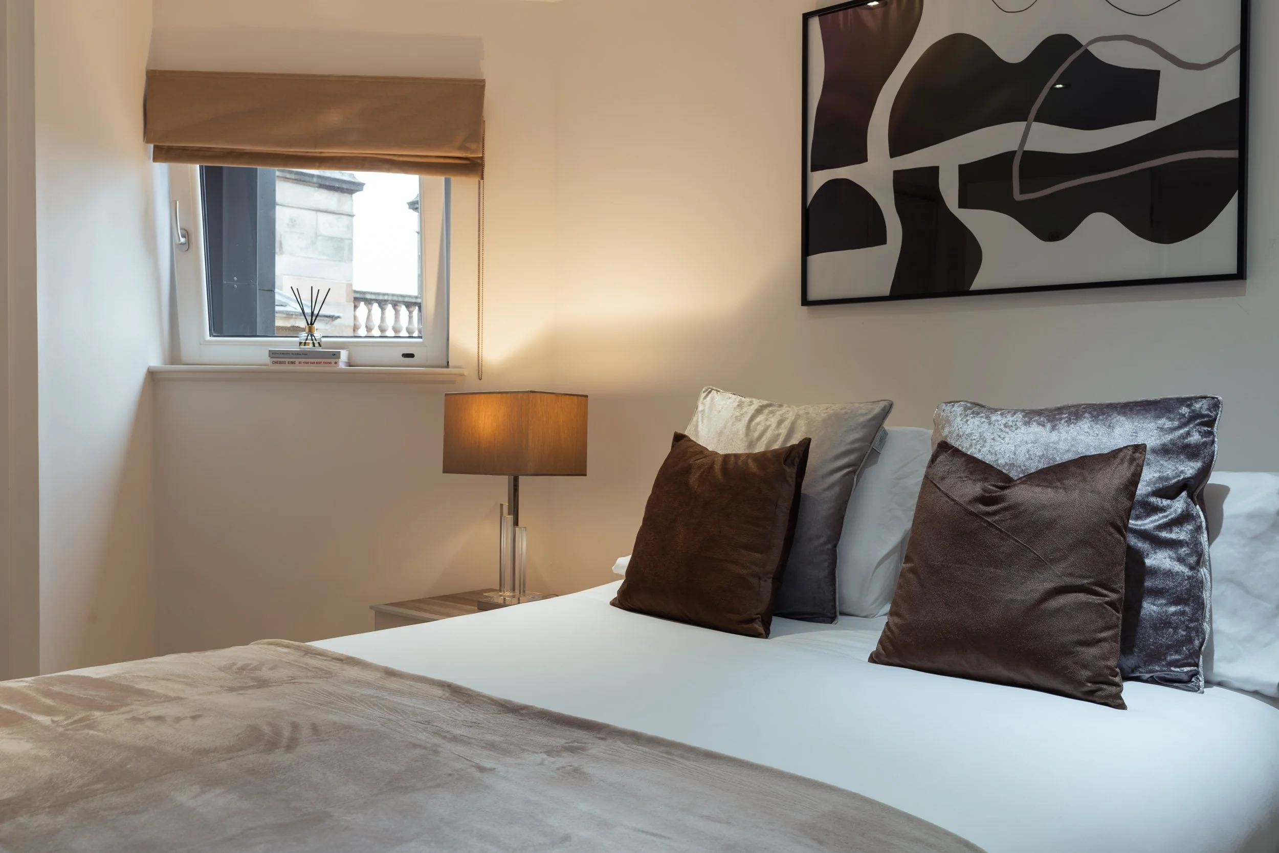 Close-up of a neatly made bed with decorative pillows in a modern bedroom. A window with a beige shade and a ledge featuring books and a reed diffuser. A beige rug covers part of the bed, and a bedside table with a table lamp is visible. Abstract art