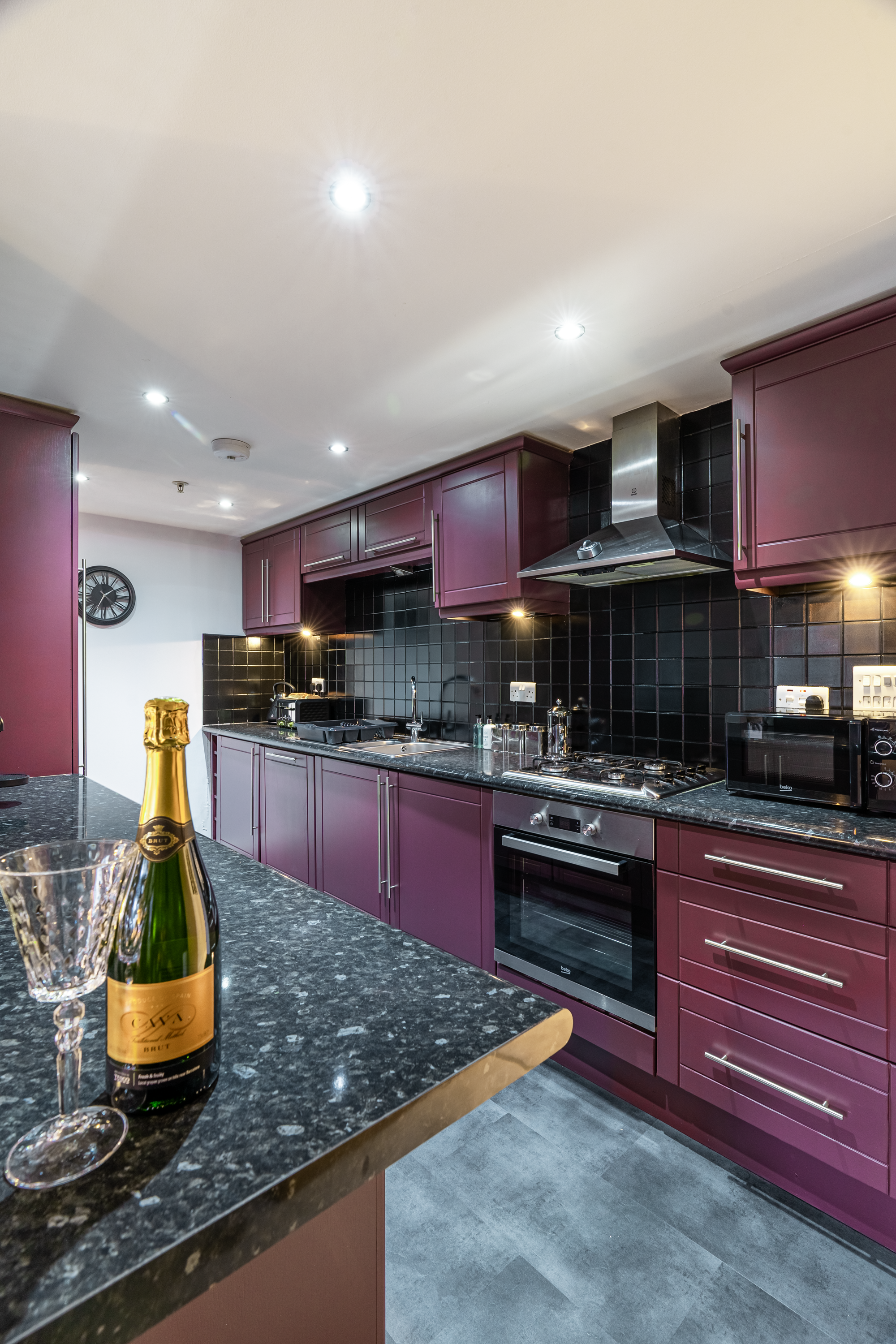 Modern kitchen with dark purple cabinets, black tiled backsplash, stainless steel oven and range hood, black countertop, and a champagne bottle with glass on a black speckled counter.