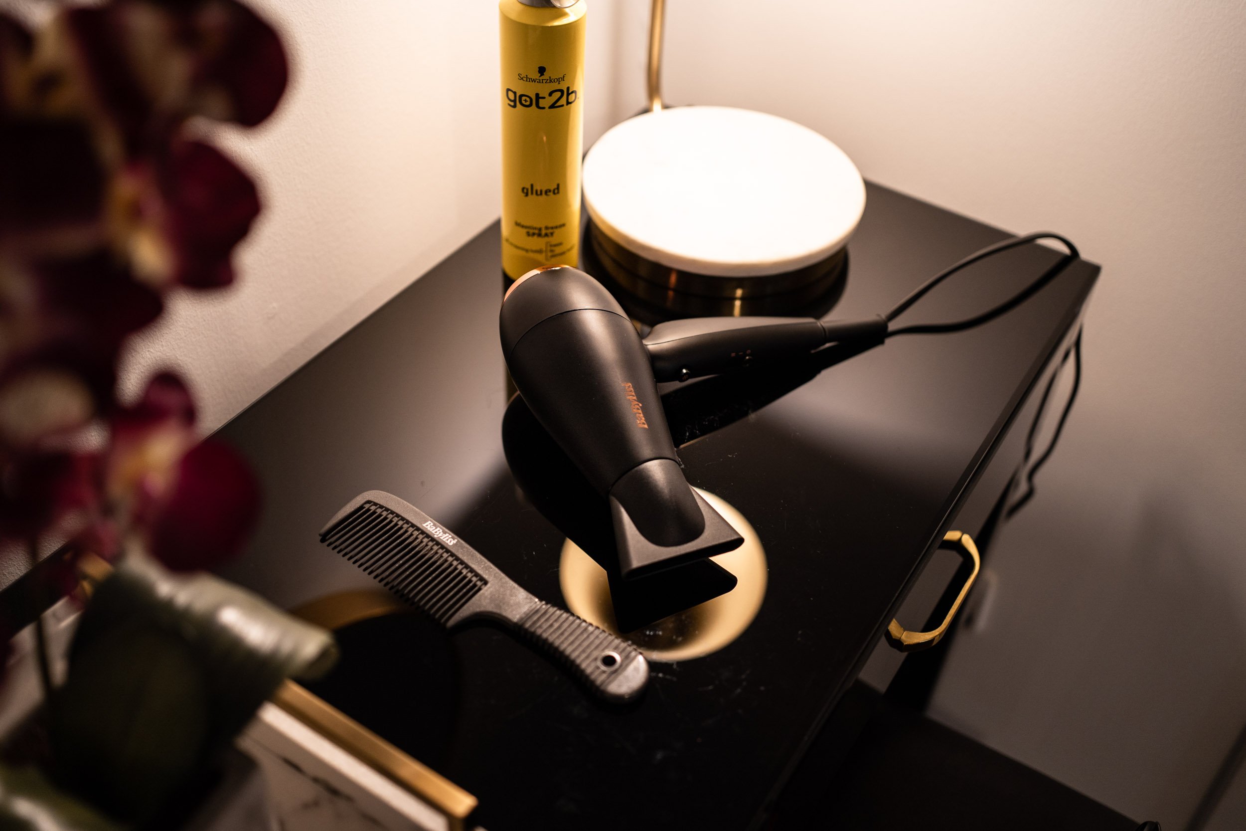 Black hairdryer, black comb, a yellow spray can, a round white lamp, and a black tray on a black table against a white wall.